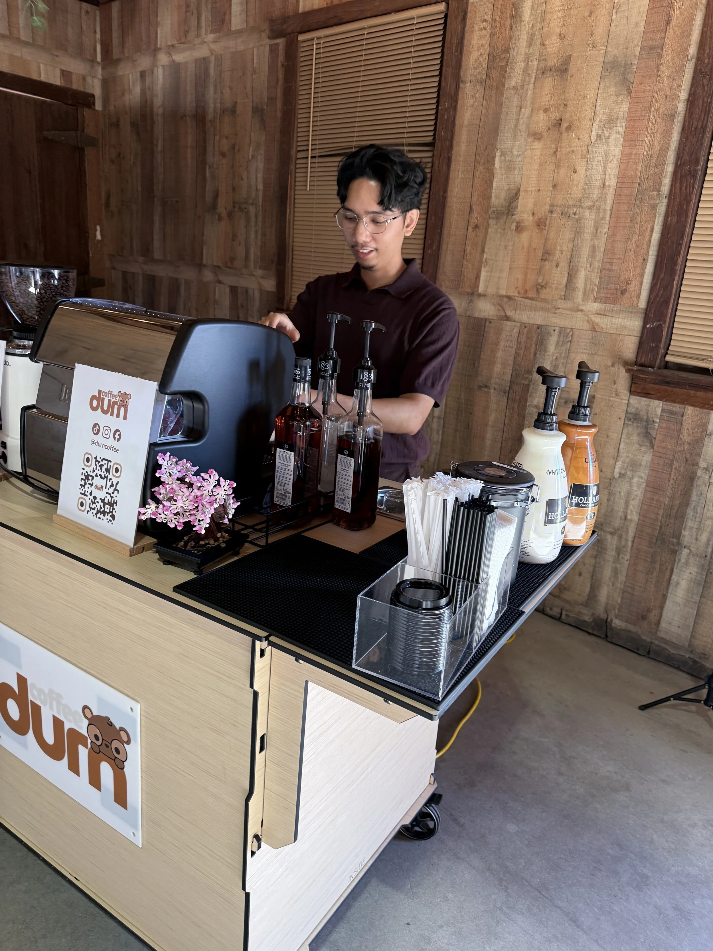 A person with glasses standing behind a coffee cart with a bear logo, preparing drinks inside a wooden room. The cart has bottles of syrup, a coffee machine, and various utensils.
