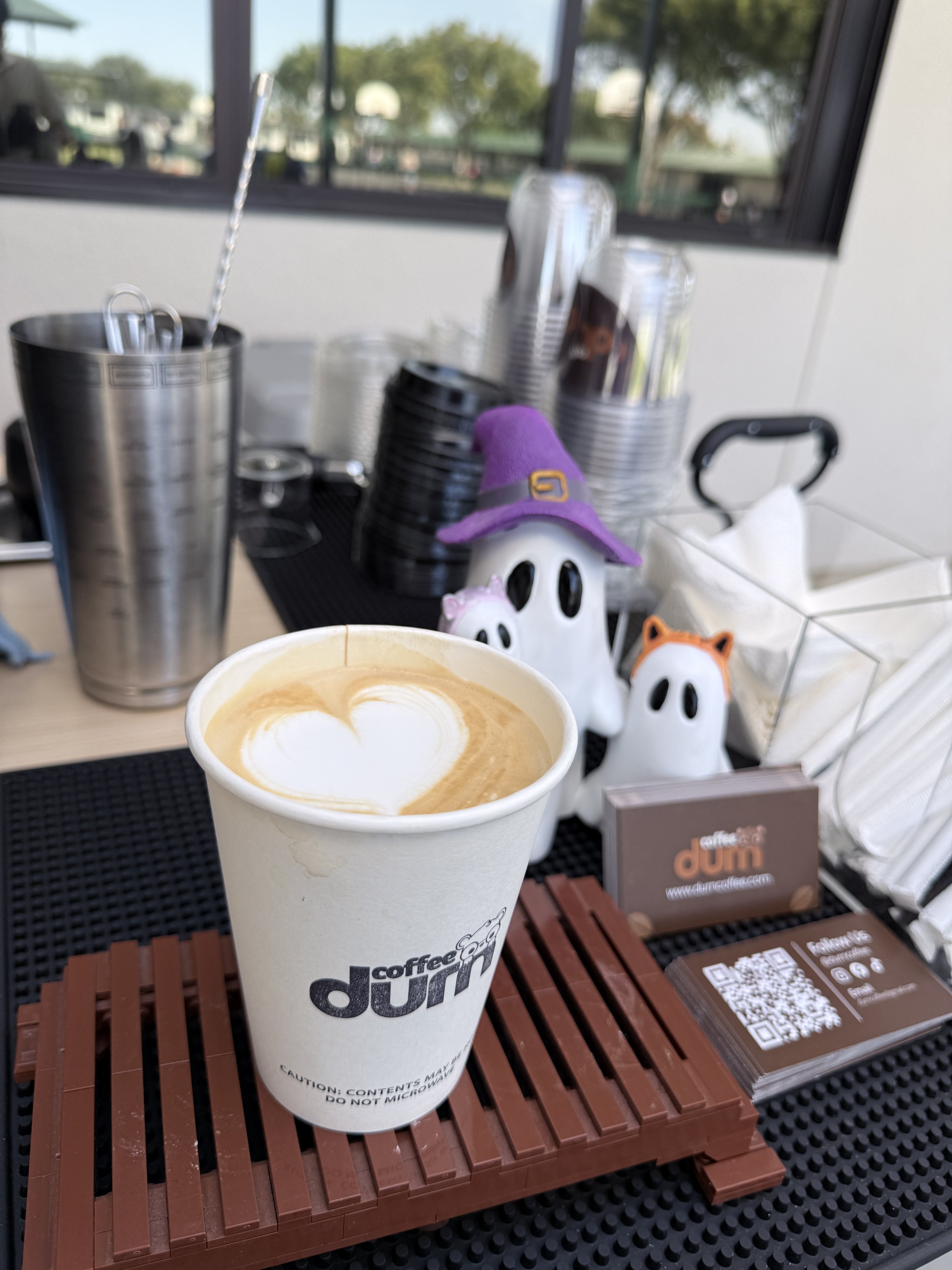 A paper cup of coffee with foam art on top, placed on a wooden table at a cafe. In the background, there are black disposable coffee cups, a small skeleton figurine wearing a purple witch hat, and some business cards and QR codes.