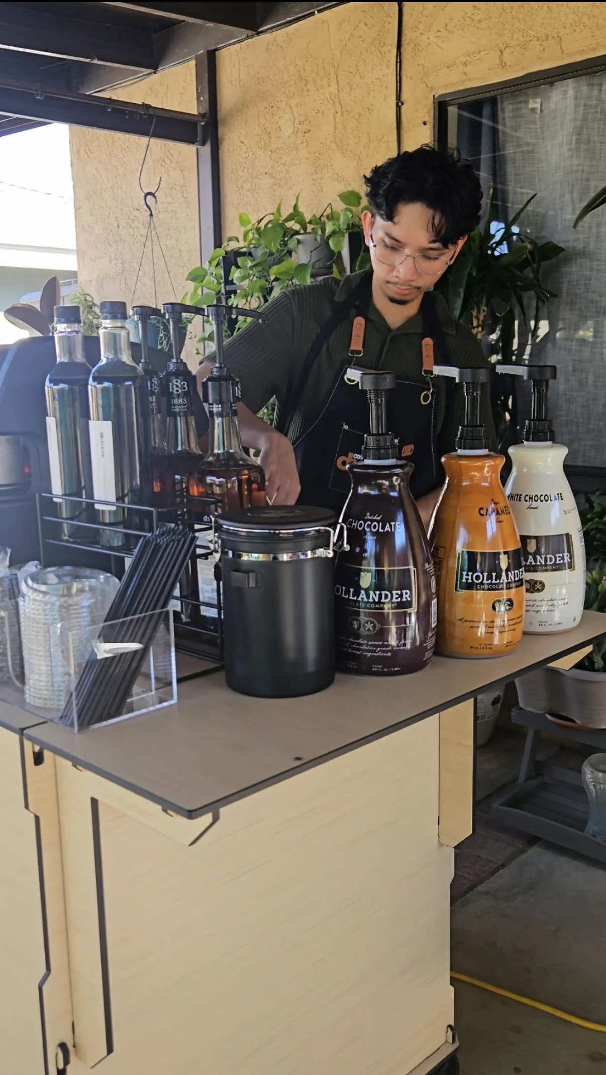 Barista preparing coffee behind a counter with bottles of chocolate syrup, a black container, and clear cups, with plants and a window in the background.