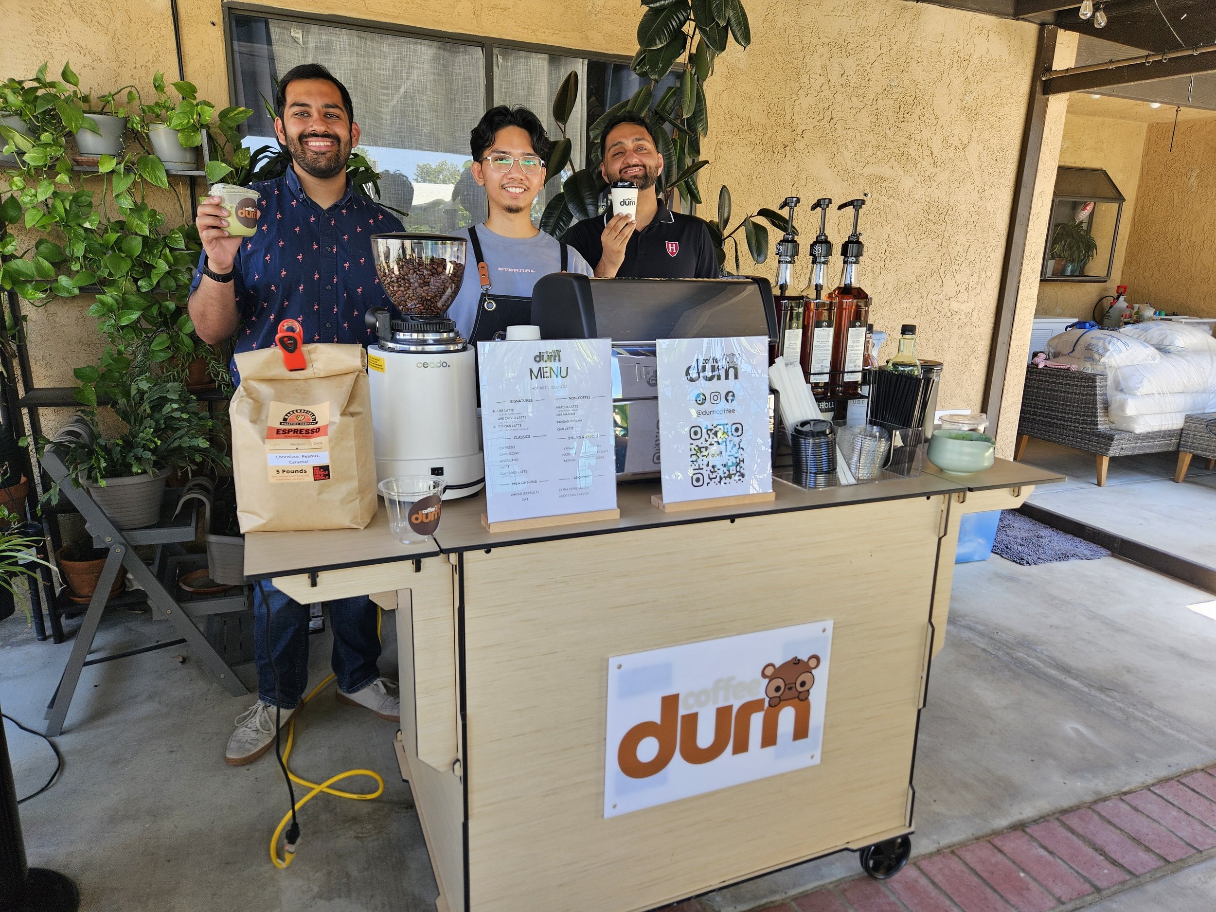 Three men behind a coffee stand smiling and holding coffee cups, with coffee beans and coffee supplies on display, outdoors under a covered patio.