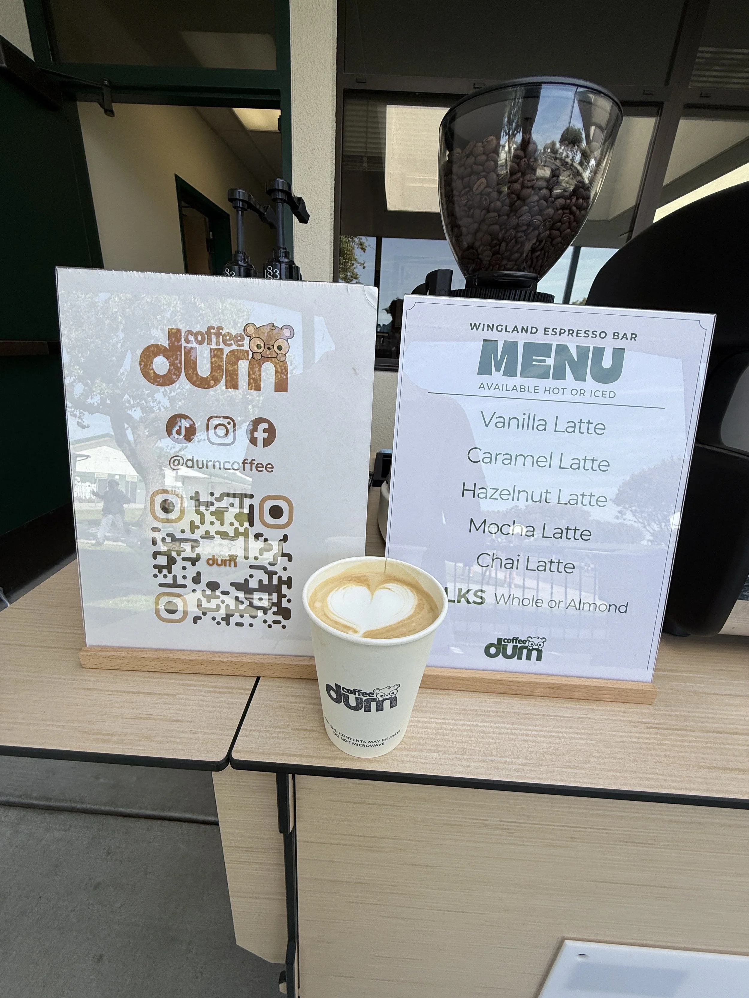 A coffee shop display with a cup of latte and latte art, a menu sign listing various latte options, and a QR code sign with social media handles, set on a wooden table outside the shop.