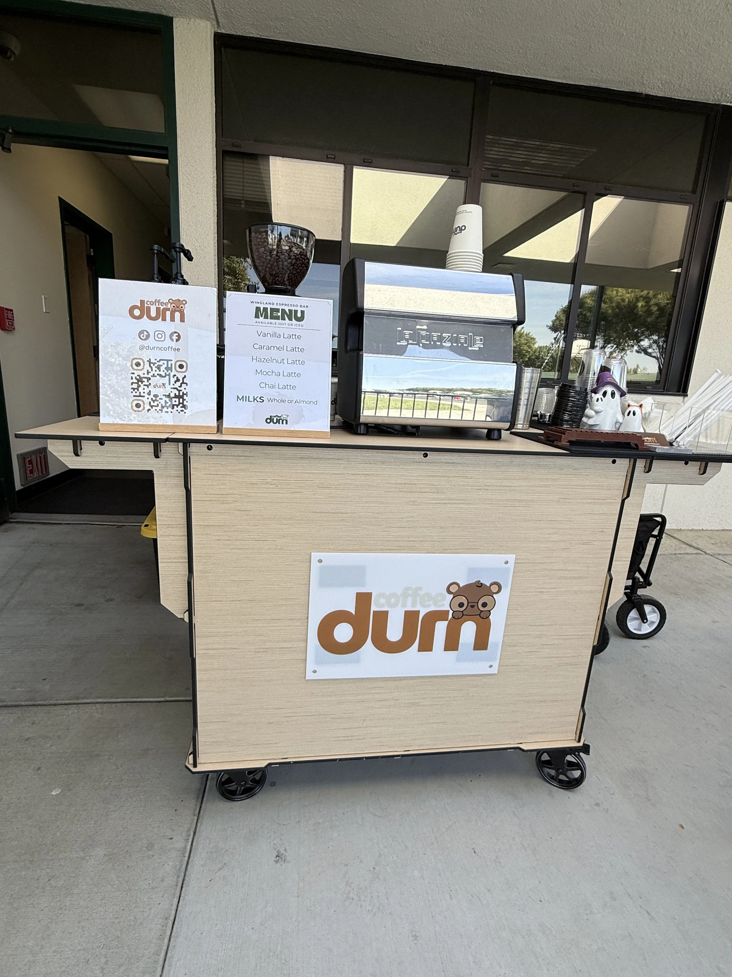 A wooden cart with a sign that says 'coffee dumi' and features a cartoon bear face. The cart has a large coffee espresso machine, a clear container of coffee beans, and a menu listing various coffee drinks such as vanilla latte, caramel latte, hazelnut latte, mocha latte, and chai latte, with milk options of whole or almond. There are disposable cups, paper cups, and Halloween decorations on the cart, with a window and trees in the background.