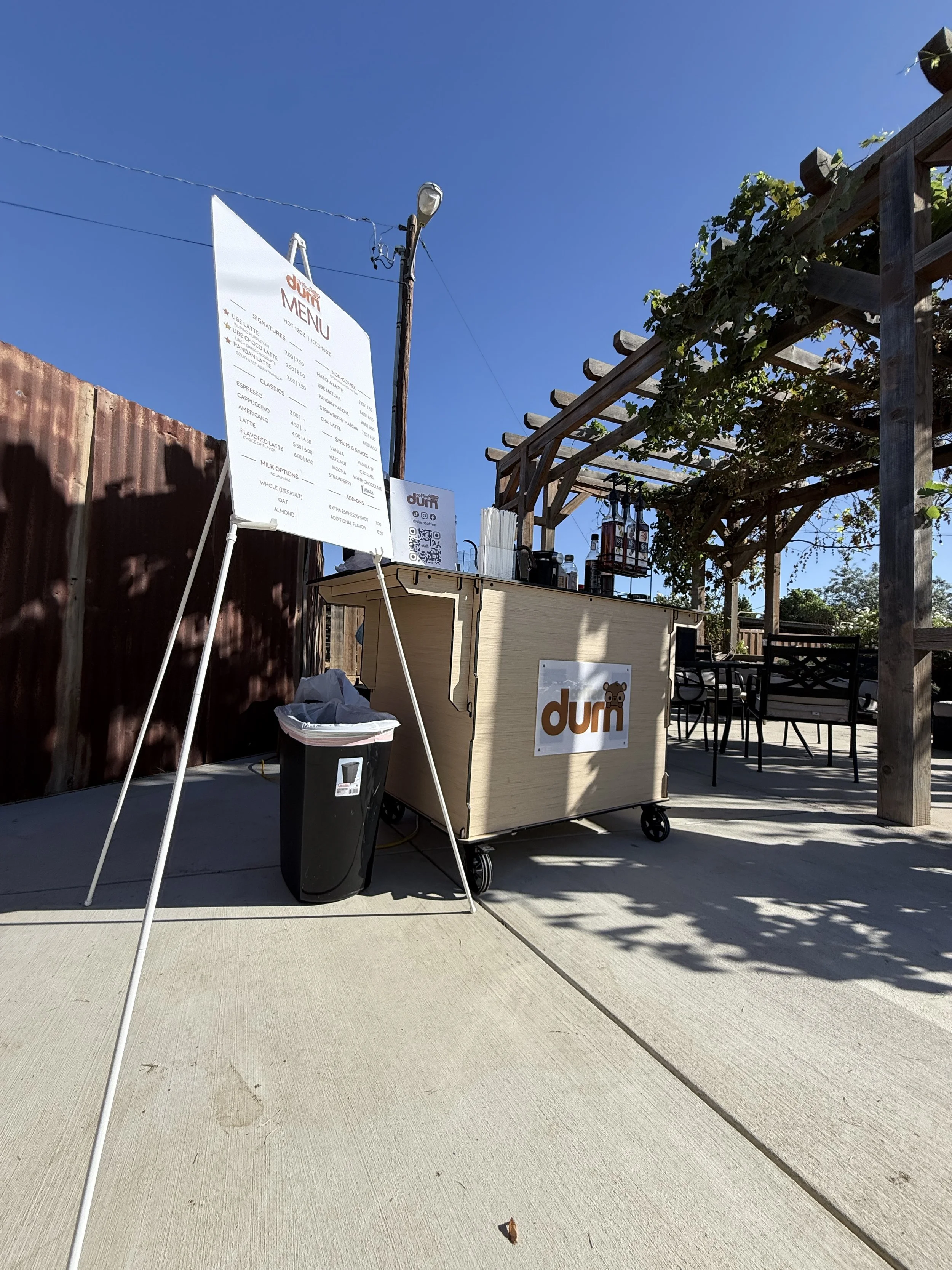 Outdoor scene with a wooden bar cart labeled 'durn', a large menu sign, and a pergola with vines, under a clear blue sky.