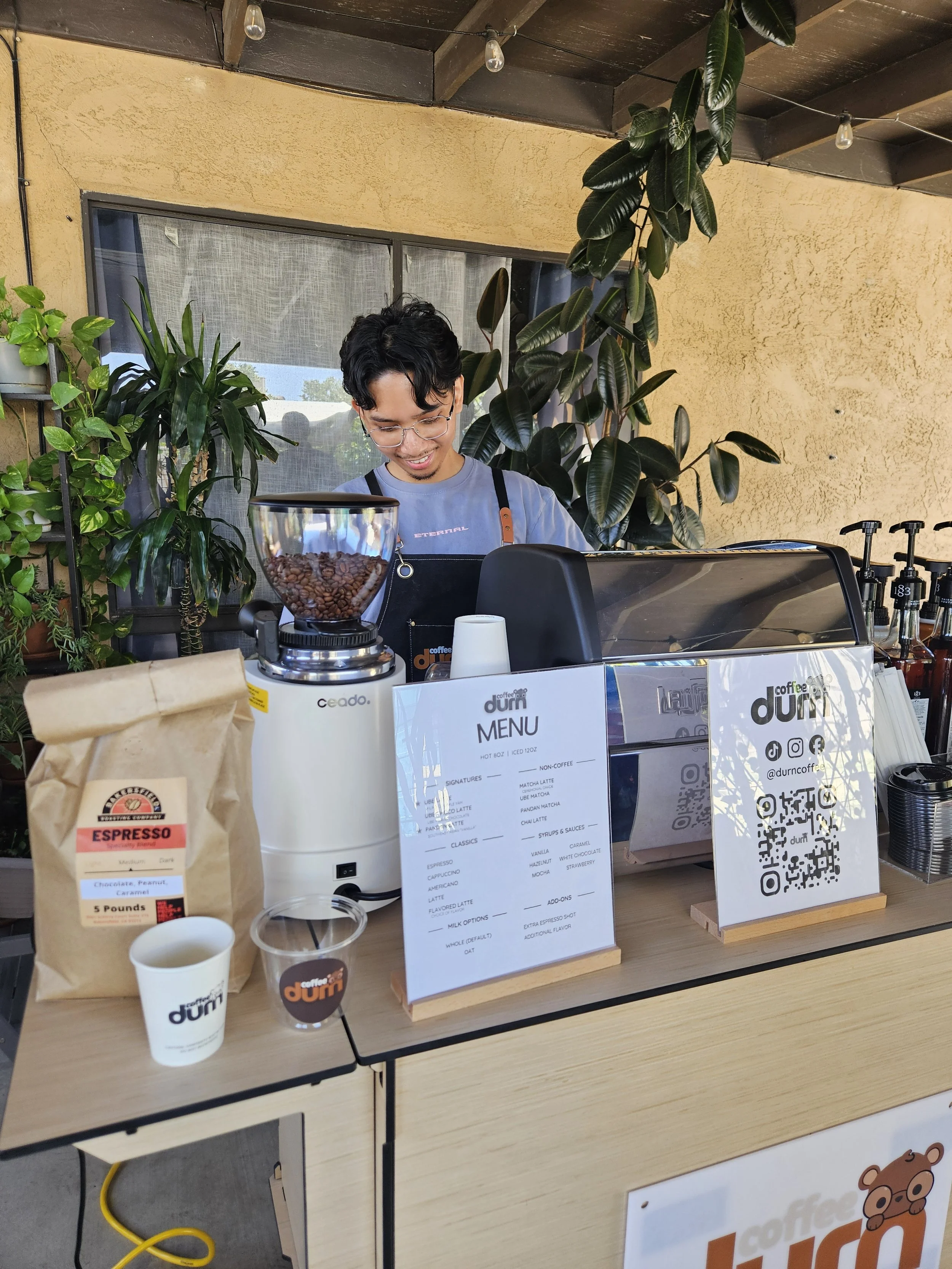 A barista inside a coffee shop, smiling while working behind the counter, with green plants on the wall, large window, coffee equipment, and a menu on the counter.