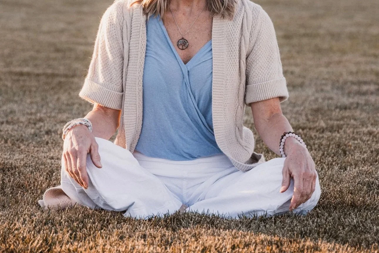 A woman sitting cross-legged on grass, with a large sun pendant necklace, wearing a blue top, beige cardigan, and white pants, during sunset or sunrise.
