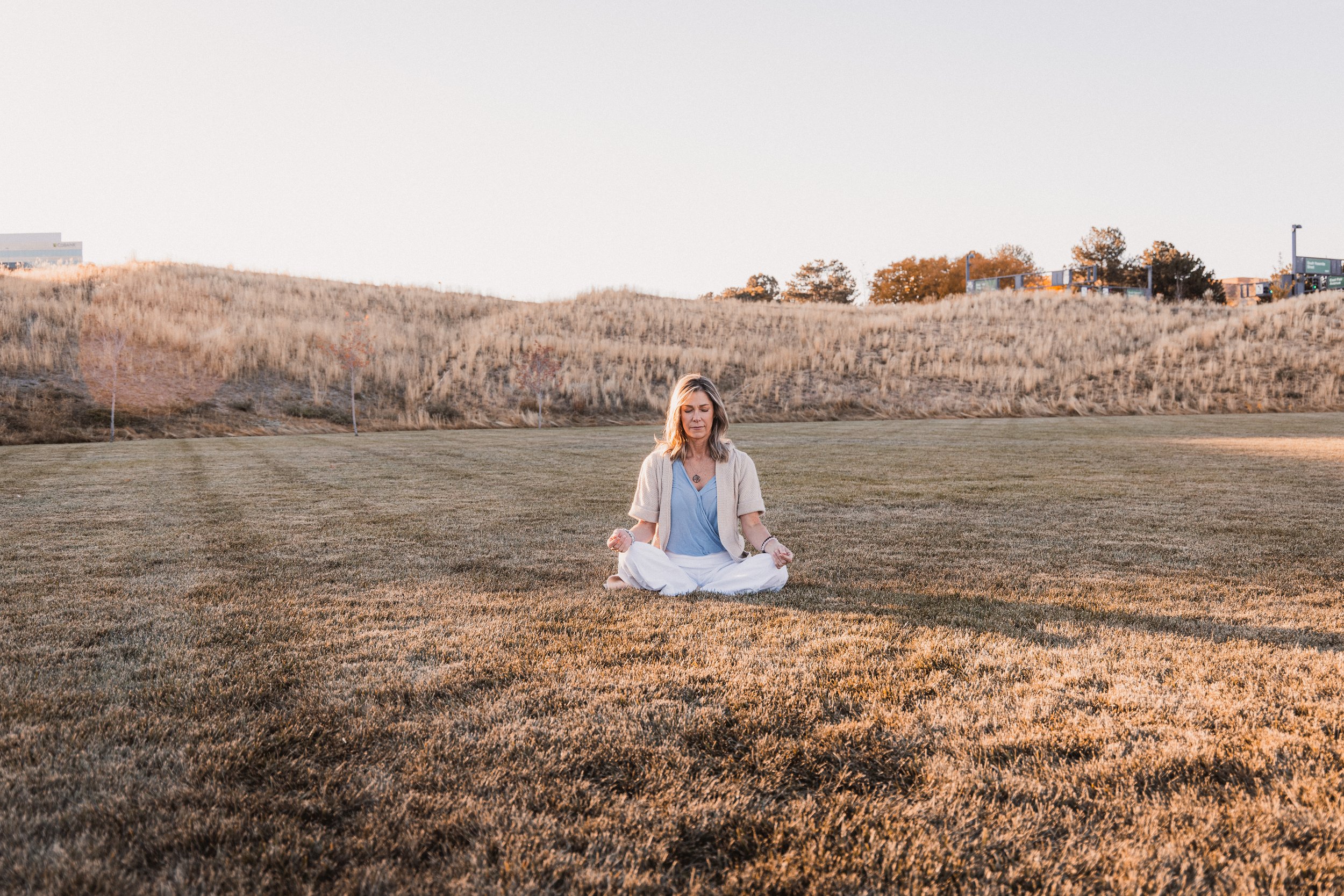 A woman sitting cross-legged on a grassy field during sunset, practicing meditation with closed eyes and relaxed expression.