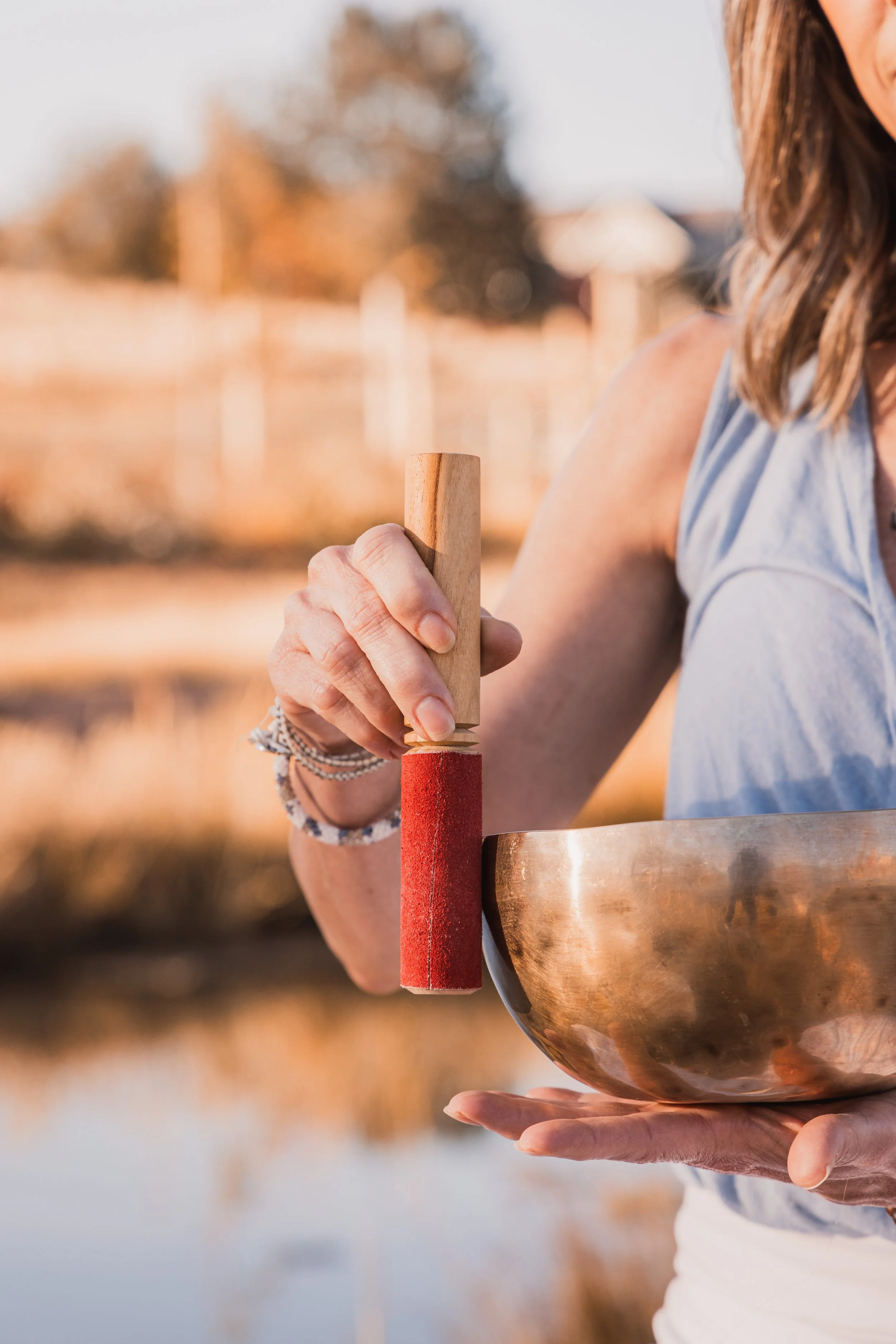 A woman holding a singing bowl with one hand and a wooden mallet with the other, outdoors during sunset.