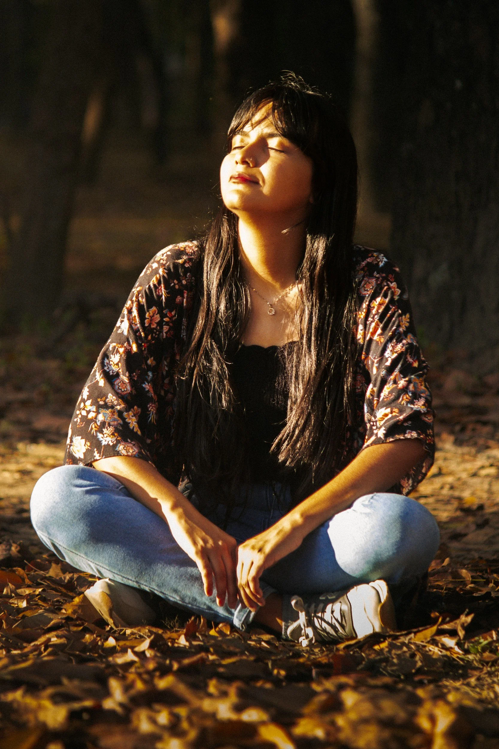 A young woman with long dark hair sitting cross-legged on autumn leaves in a forest, basking in sunlight with her eyes closed.