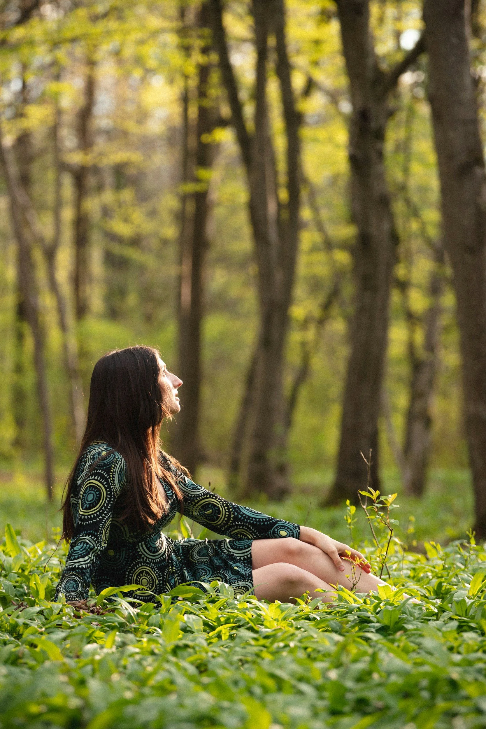 A woman sits on the ground in a lush green forest, surrounded by foliage and trees, looking up peacefully.