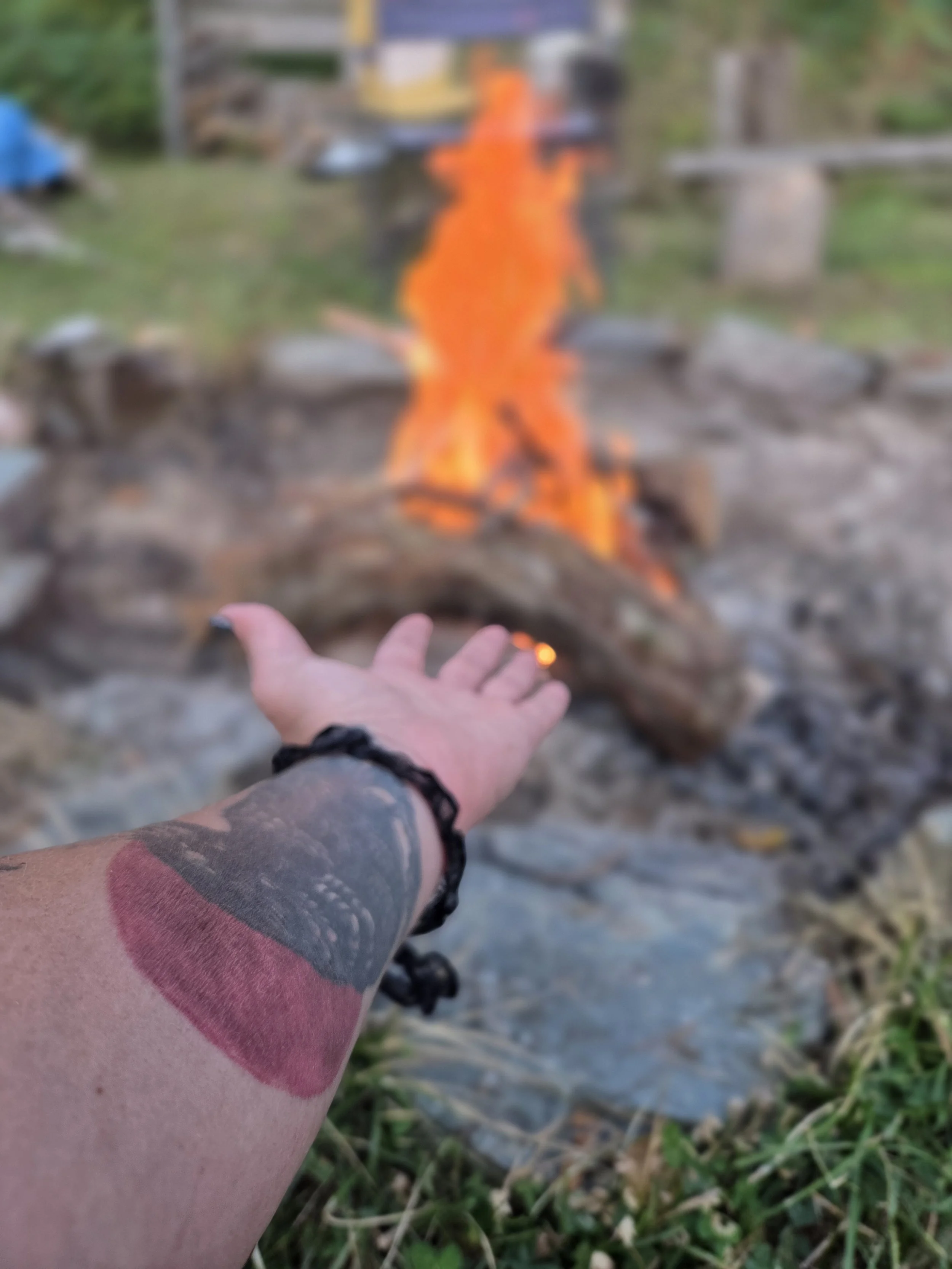 Person with tattooed arm reaching out towards a campfire with orange flames, surrounded by rocks and grass.