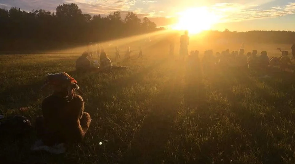 People sitting and standing in a grassy field at sunset, with sunlight streaming through trees and haze in the air.