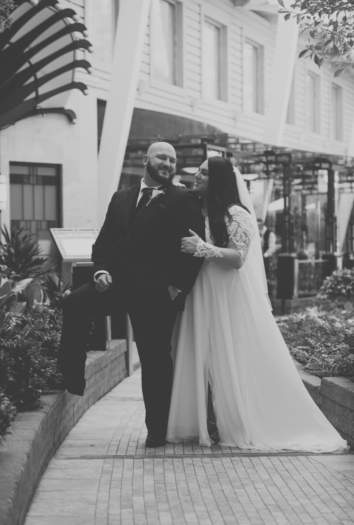 A black and white photo of a bride and groom standing outdoors, smiling, with greenery and a modern building in the background. The bride is wearing a long white wedding dress with lace sleeves, and the groom is in a dark suit with a tie. The groom is sitting on a ledge with one leg crossed, and the bride is leaning towards him, holding his arm.