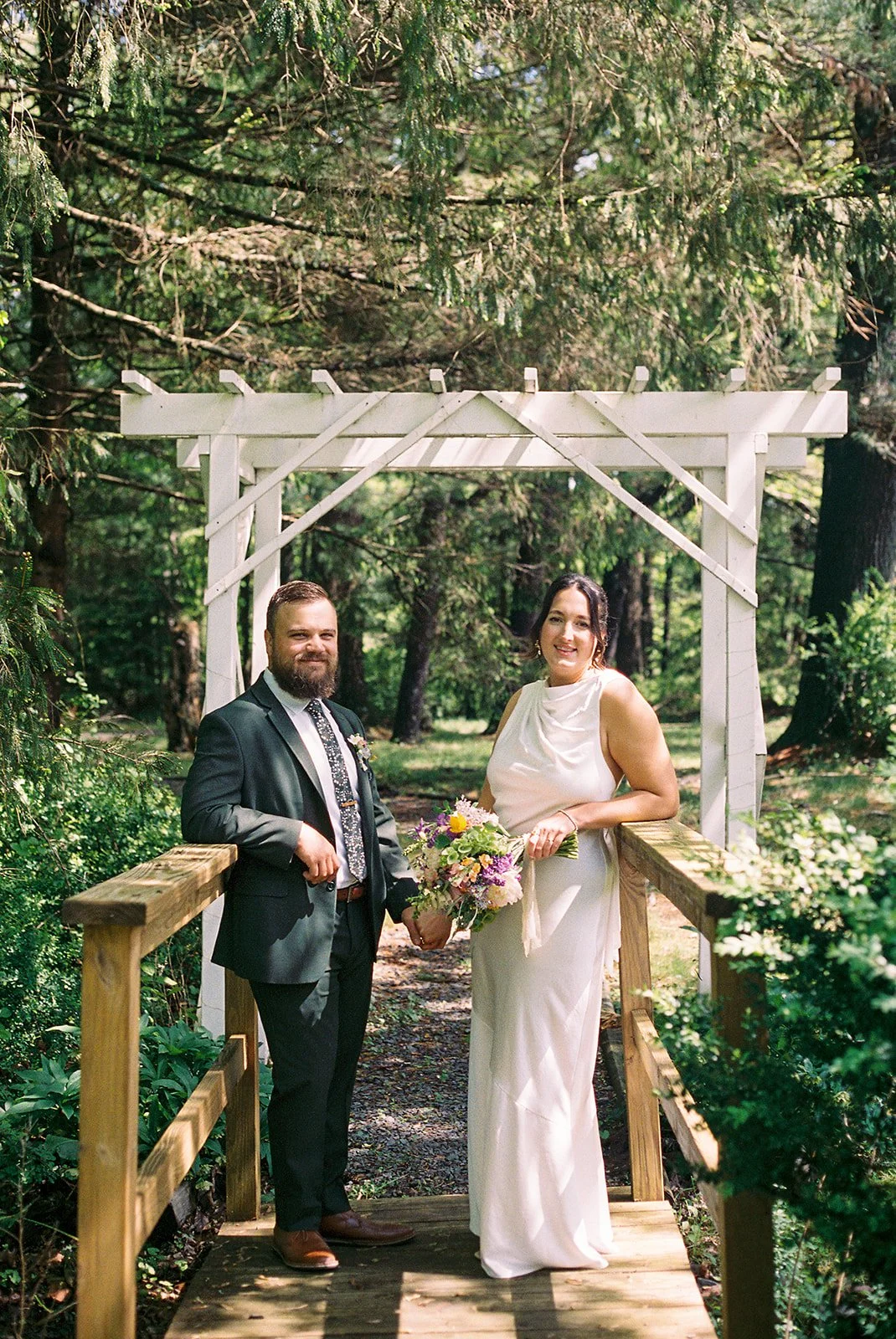 A bride and groom standing on a wooden bridge in a lush forest, smiling, with a white wooden arch behind them, the bride holding a bouquet of flowers.