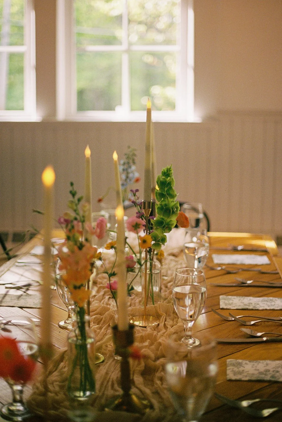 A dining table decorated for an event with candles, flowers, glasses of water, and cutlery, set in a room with sunlight coming through a window.