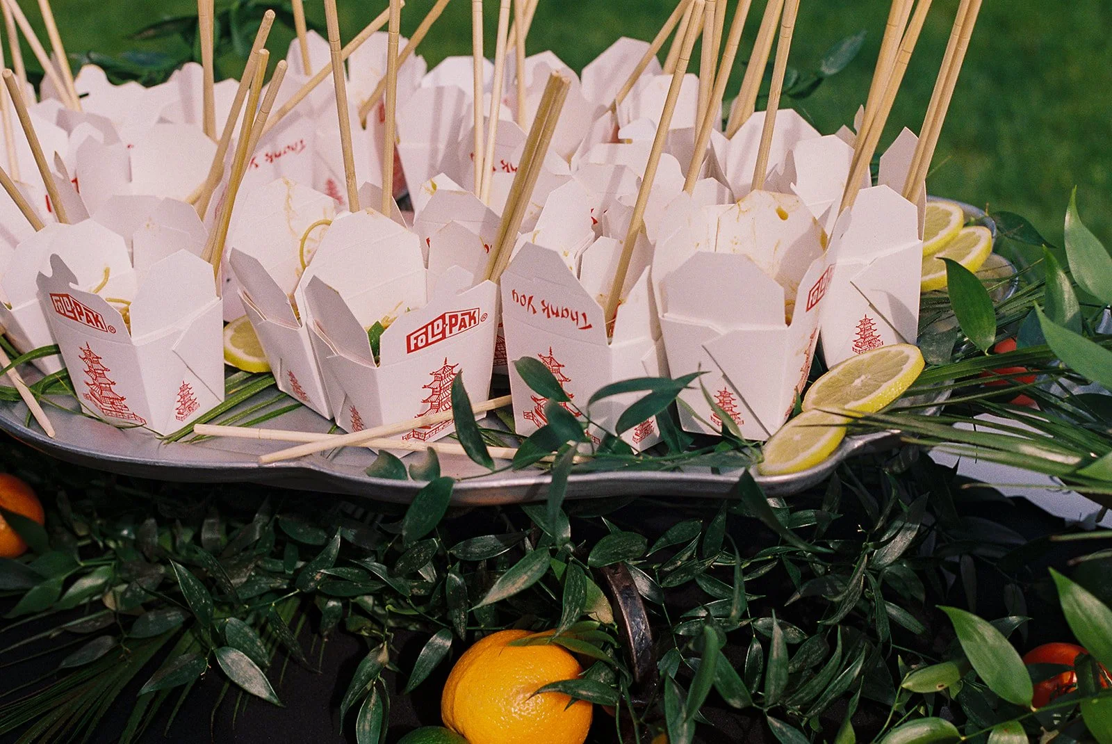 A tray with small paper food containers labeled 'FoL-PaK' filled with food, garnished with lemon slices, surrounded by green leaves and oranges.