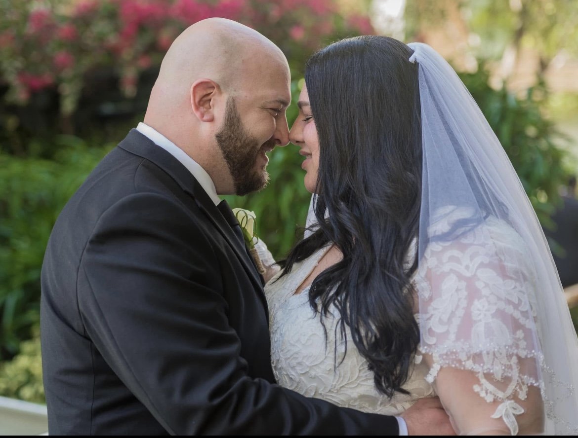 A bride and groom face each other close, touching foreheads, outdoors with greenery and pink flowers in the background.