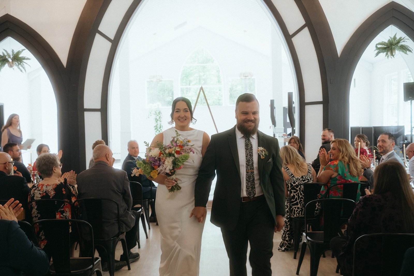 A newlywed couple walking down the aisle at their wedding reception, surrounded by seated guests clapping and smiling, inside a bright venue with large arched windows.
