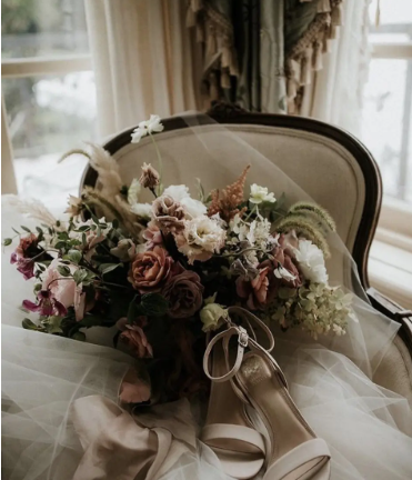 A wedding bouquet with pink, purple, and white flowers, placed on a vintage-style chair along with a pair of beige heels.