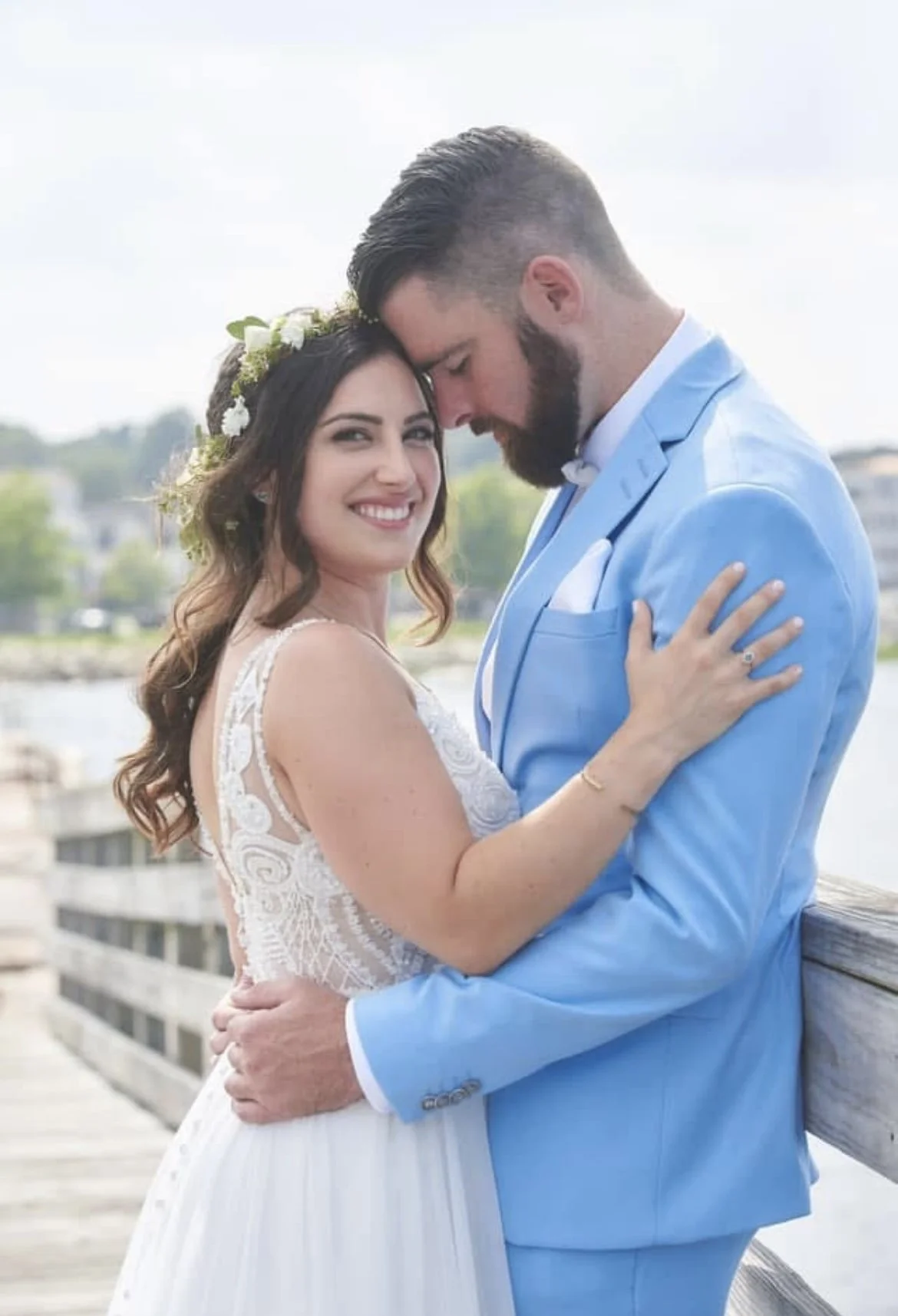 A newlywed couple stands on a wooden dock outdoors, embracing with their foreheads touching. The bride is smiling at the camera, wearing a white wedding dress with lace details and a floral crown. The groom is dressed in a light blue suit with a white shirt and bow tie, looking down at her. The background features a body of water, trees, and a partly cloudy sky.