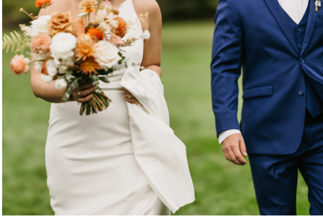 Bride holding a bouquet of flowers while walking with the groom on a grassy field.