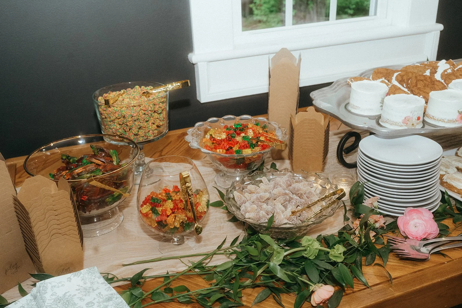 Decorative table with bowls of colorful candies, cookies, and small cakes, surrounded by a green garland and pink flowers.