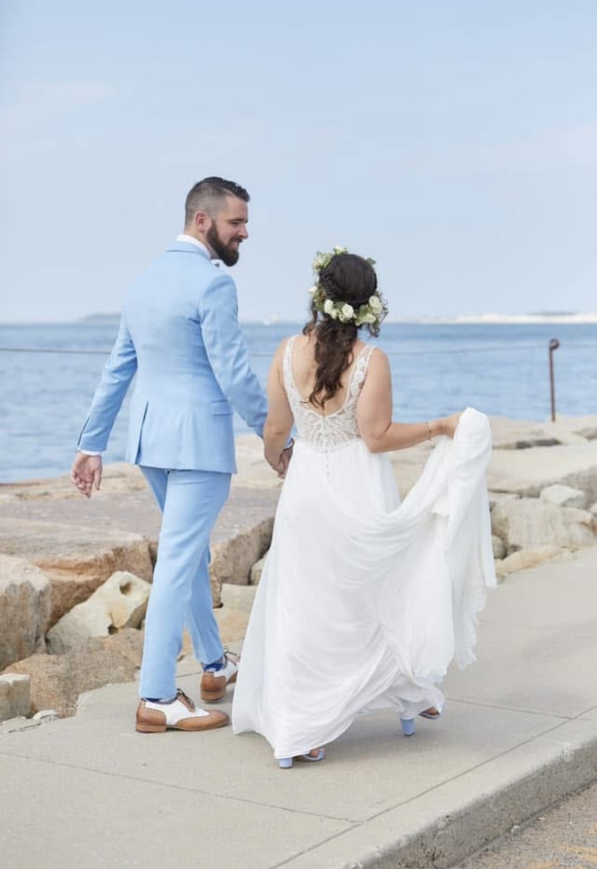 A couple dressed in wedding attire walking along a waterfront, holding hands. The woman wears a white wedding dress with a floral crown, and the man wears a light blue suit.