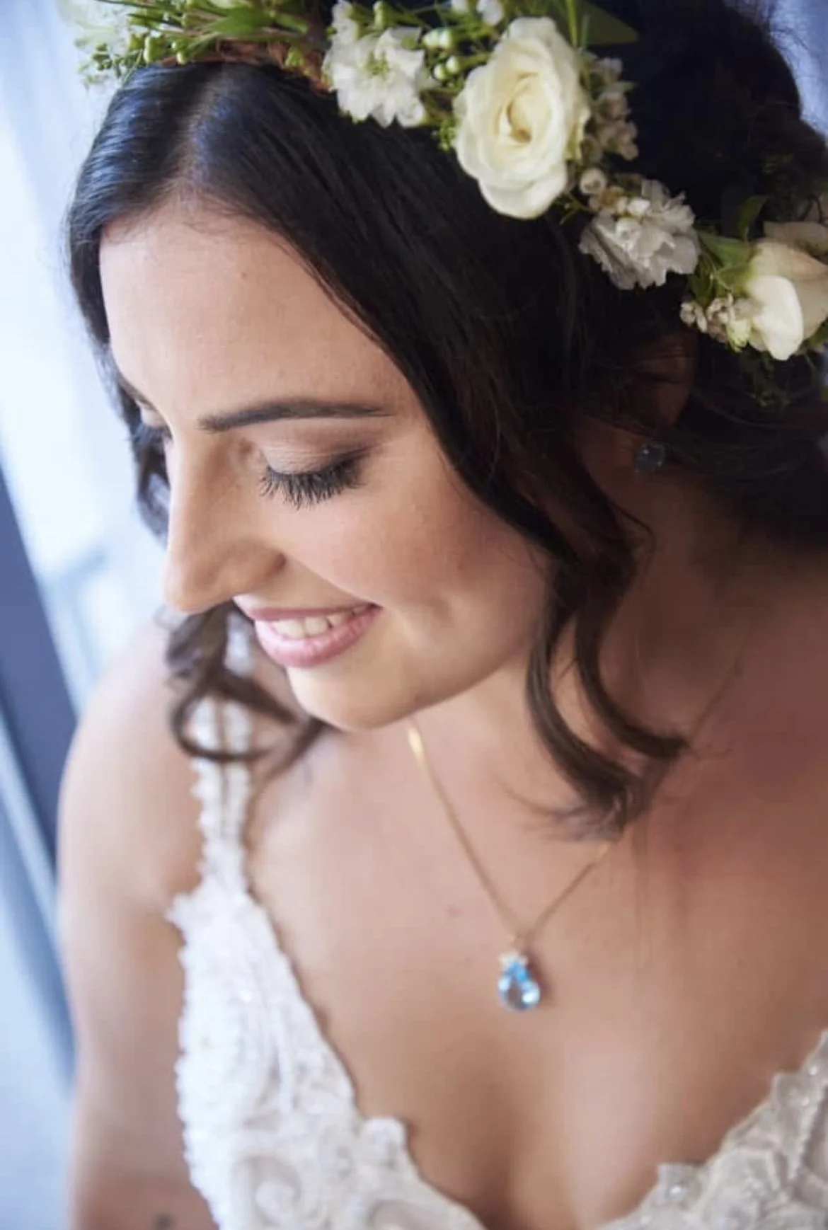Close-up of a smiling bride with dark hair wearing a white lace wedding dress and a floral crown, featuring white roses and greenery, with a gold necklace and matching earrings, looking down.