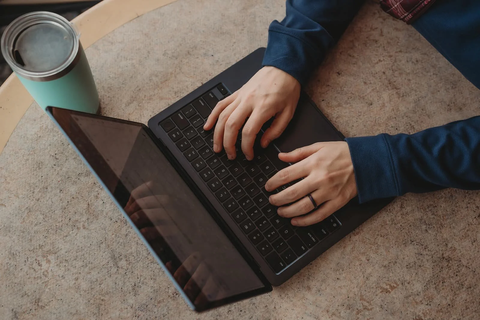 Person typing on a laptop with a beverage container nearby on a beige surface.