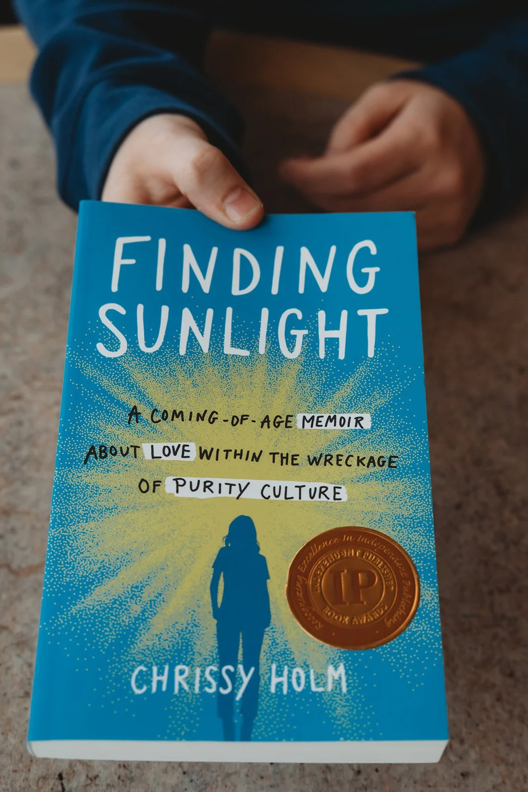 Person holding a book titled "Finding Sunlight" by Chrissy Holm, featuring a blue cover with a silhouette of a person and a gold medal emblem.