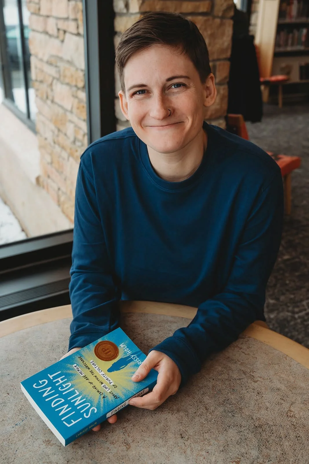 Young person with short brown hair smiling and holding a paperback book titled "Finding Sunlight" inside a room with brick wall, bookshelf, and a window showing outdoor scenery.