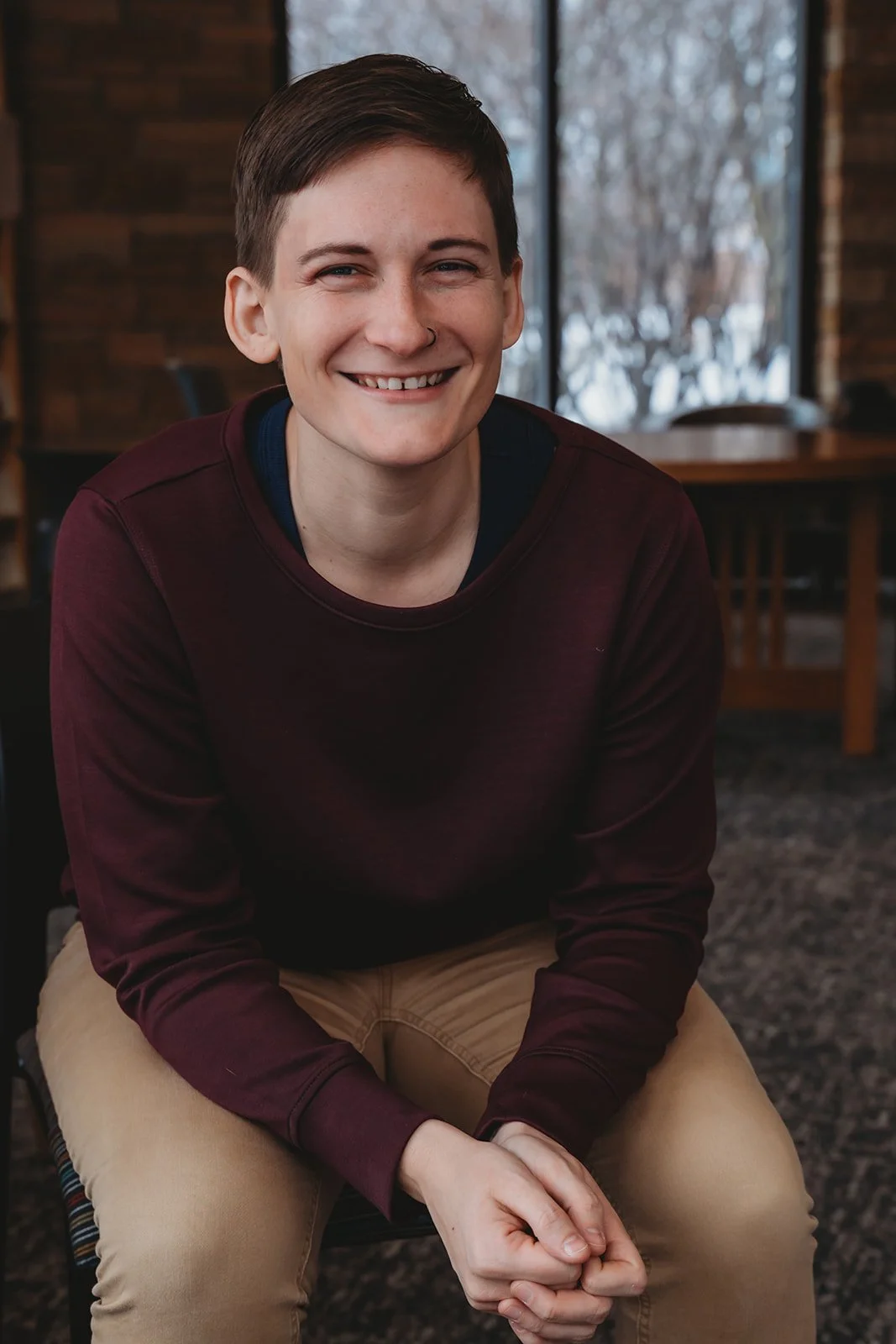 A person with short brown hair, wearing a maroon sweatshirt and beige pants, sitting indoors near a window with a smile.
