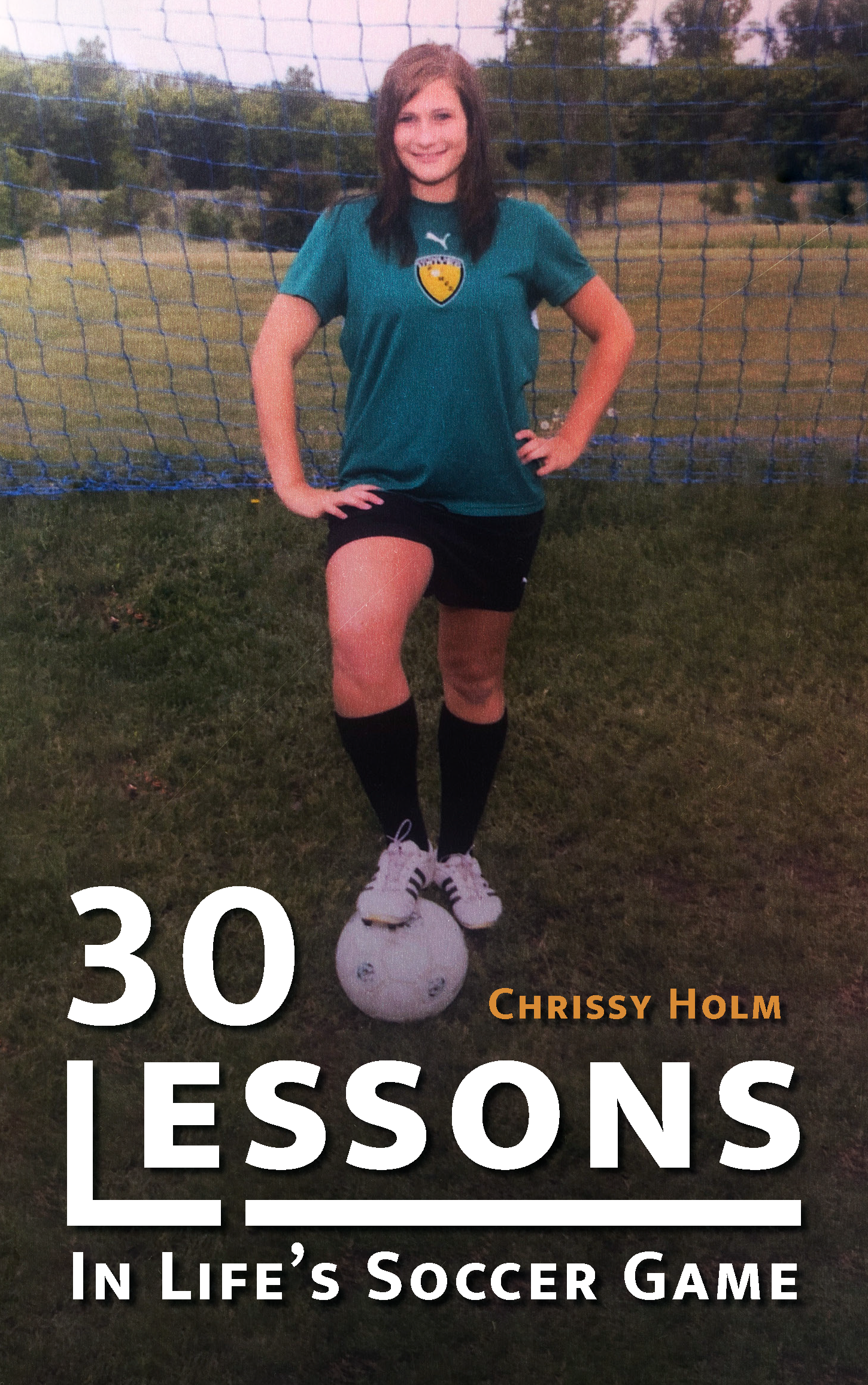 A young person with brown hair, wearing a teal soccer jersey and black shorts, stands on a soccer field with hands on hips, next to a soccer ball at feet, in front of a goal net with trees in the background.