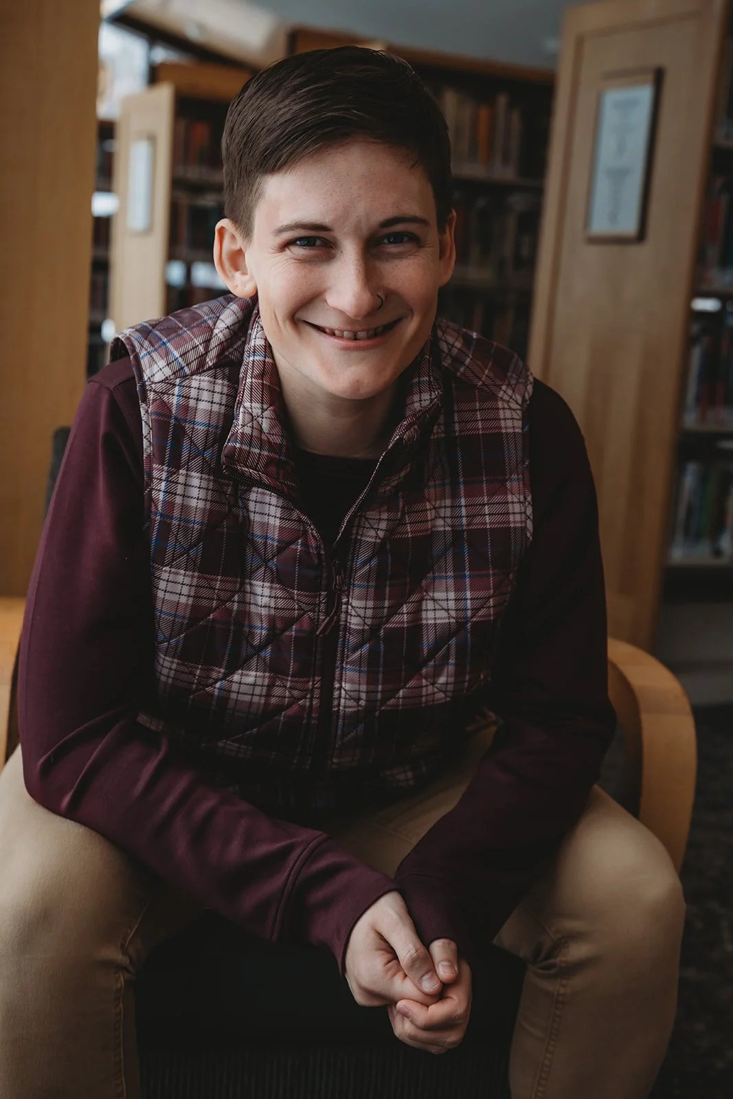 A person with short dark hair, wearing a plaid vest over a maroon long-sleeve shirt, sitting on a chair in a library with bookshelves in the background.