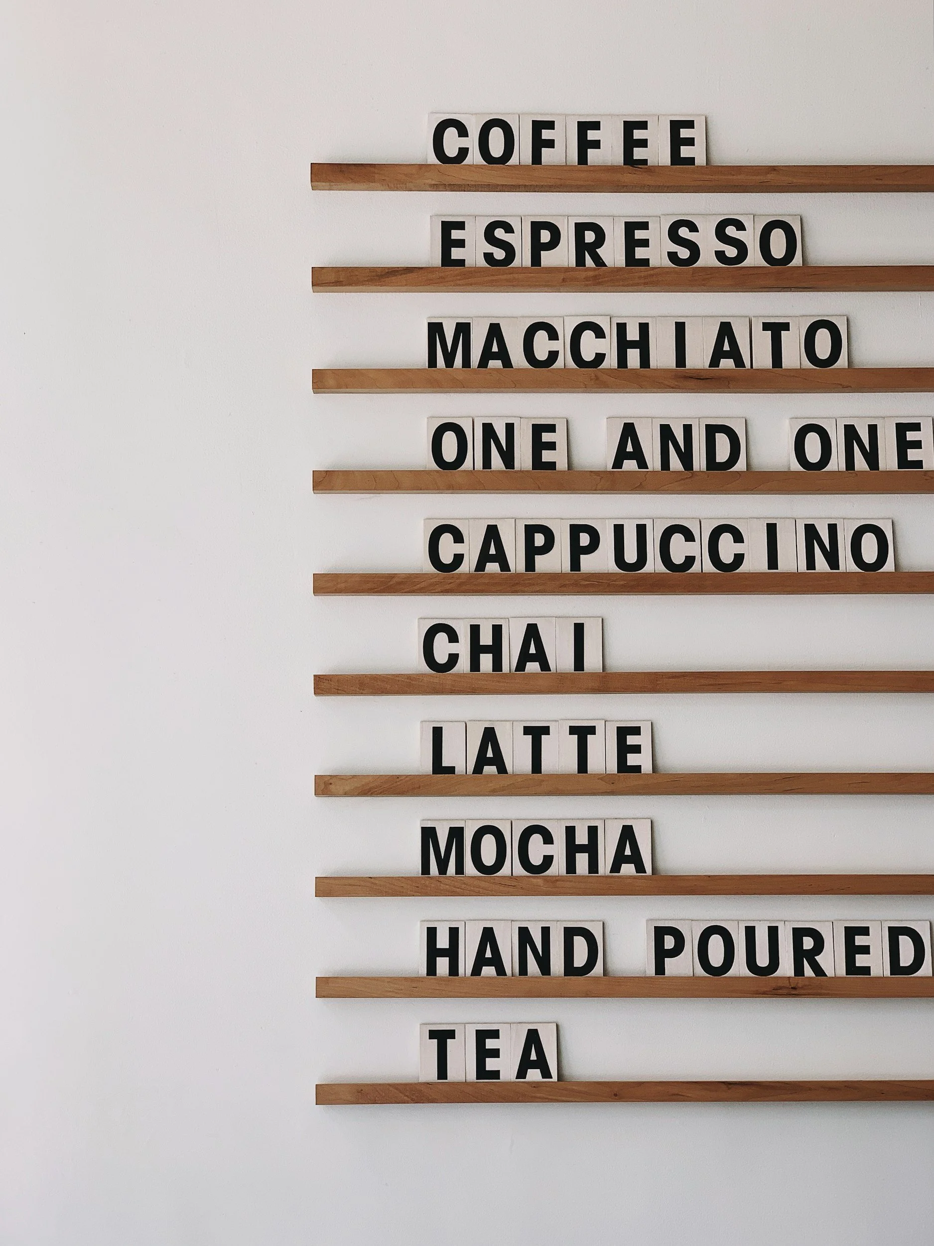 A wall-mounted menu with black letters on white tiles, listing types of coffee and related beverages in rows separated by wooden slats.