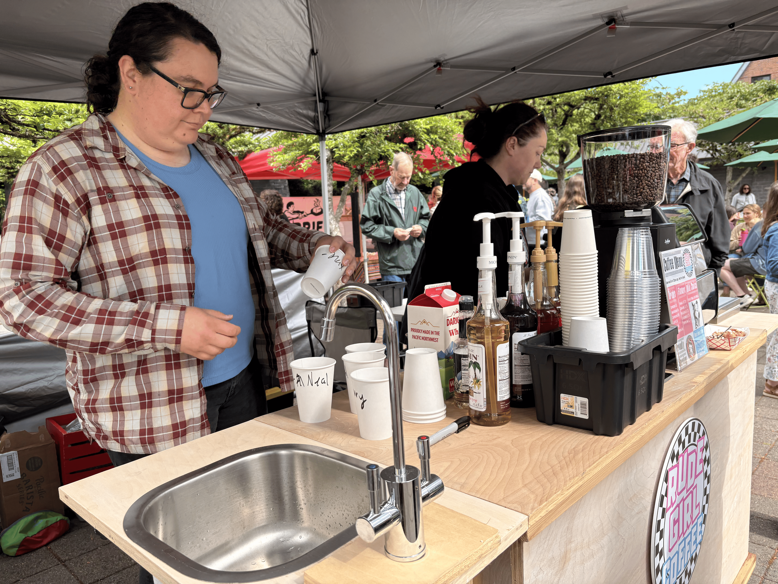 People standing behind a wooden counter at an outdoor cafe or market stall, with coffee beans and cups on the counter, under a gray canopy, surrounded by trees and other people.