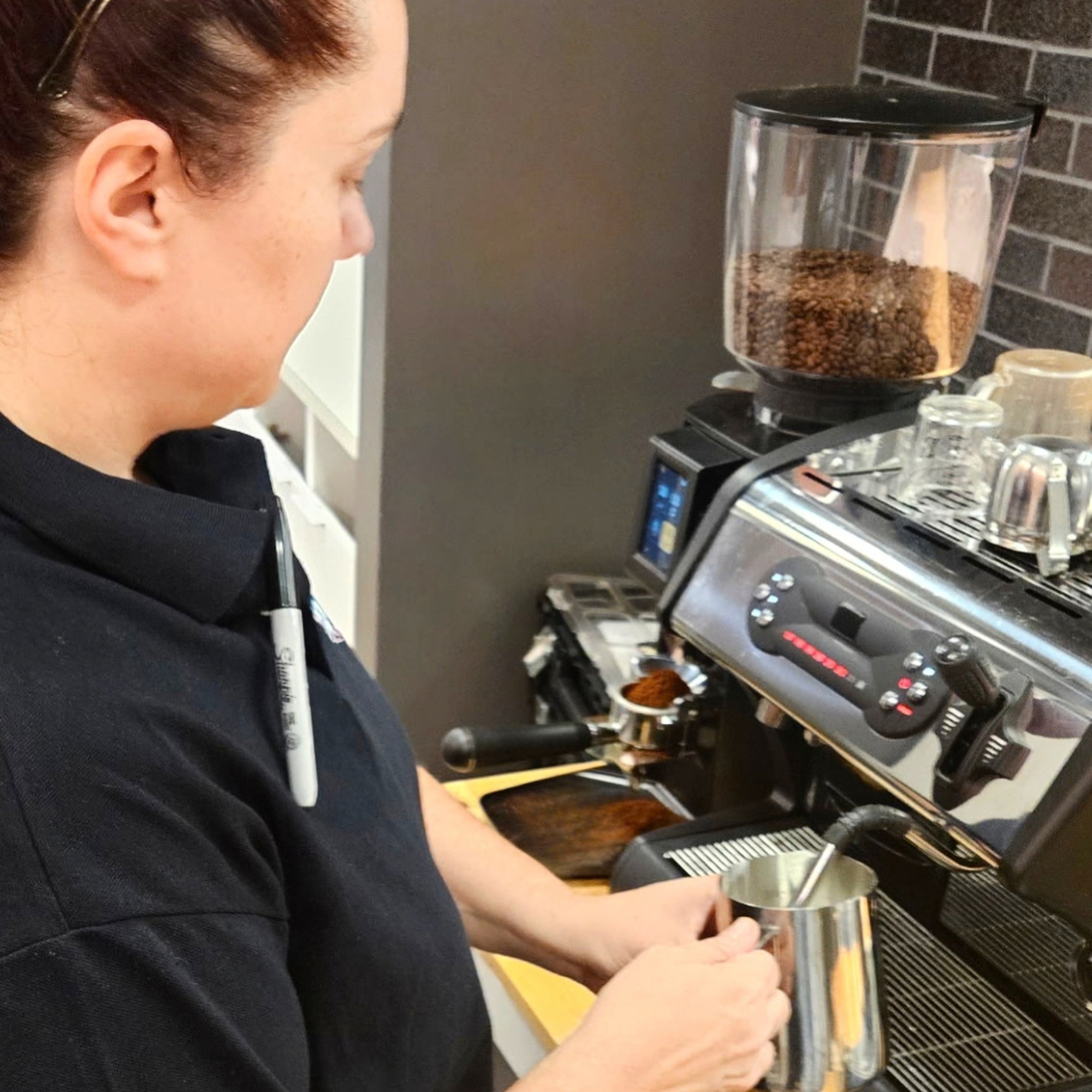 A woman with brown hair tied back, wearing glasses and a black shirt, preparing food in a kitchen. She is scooping food from a small metal bowl into a cooking appliance that appears to be a toaster oven.