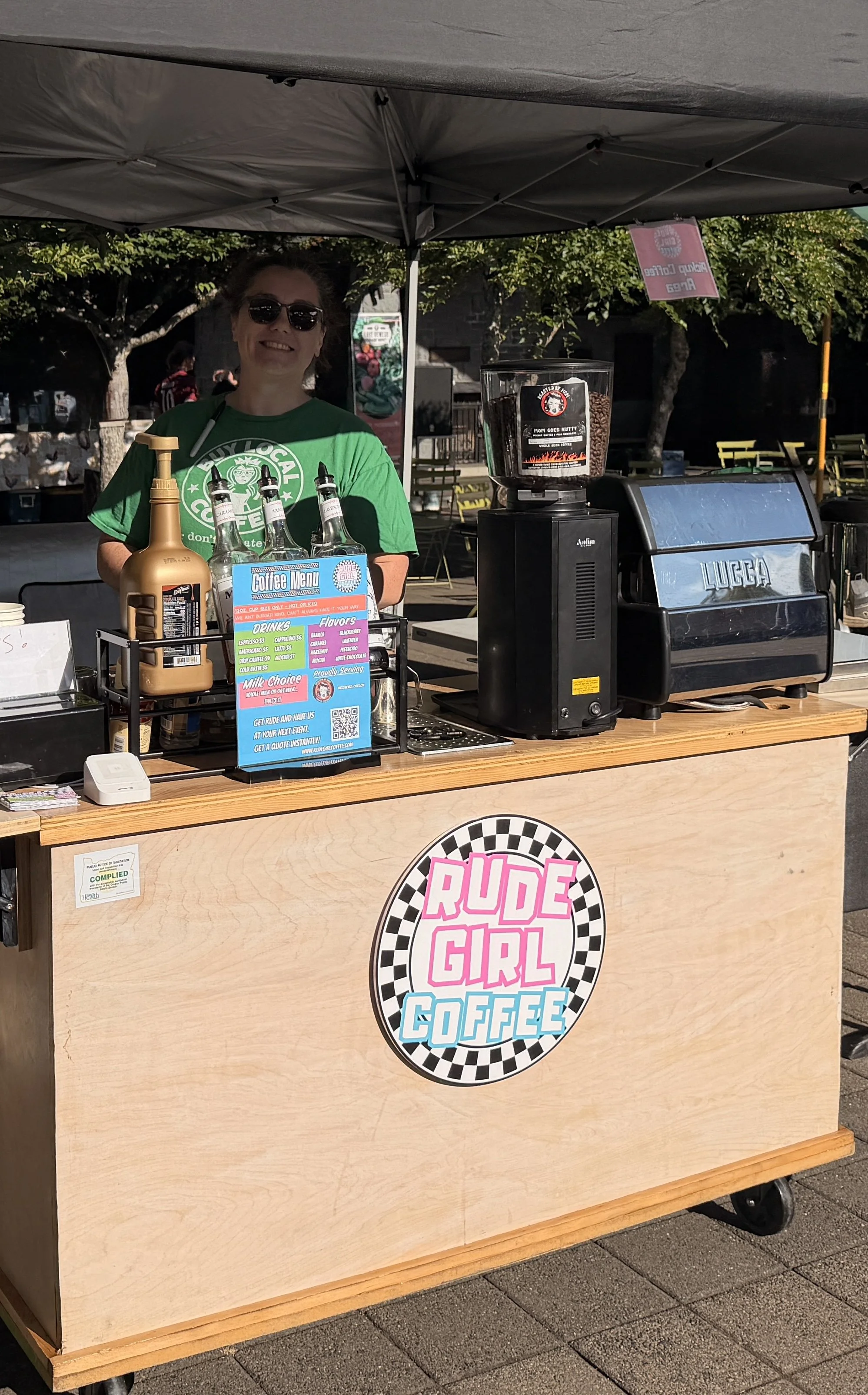 Barista behind the Rude Girl Coffee espresso cart with grinder and flavored syrups at an outdoor coffee catering setup