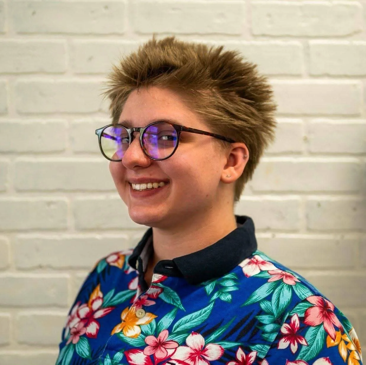 A smiling young person with short, spiky hair, wearing glasses with purple lenses and a colorful Hawaiian shirt, standing in front of a white brick wall.