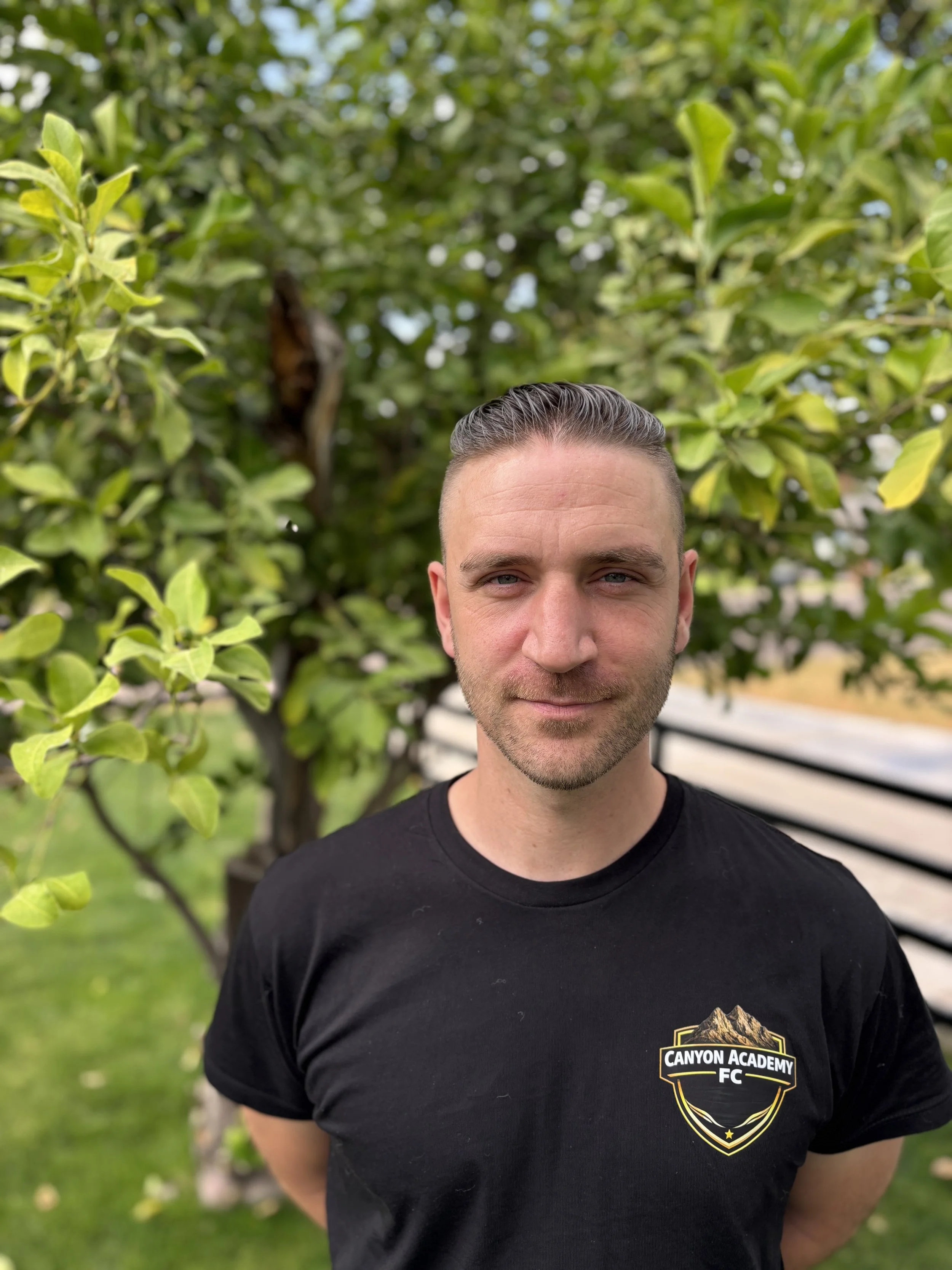 A man with short, slicked-back hair, wearing a black t-shirt with a mountain and canyon logo that says 'Canyon Academy FC,' standing outdoors in front of green foliage and a tree.