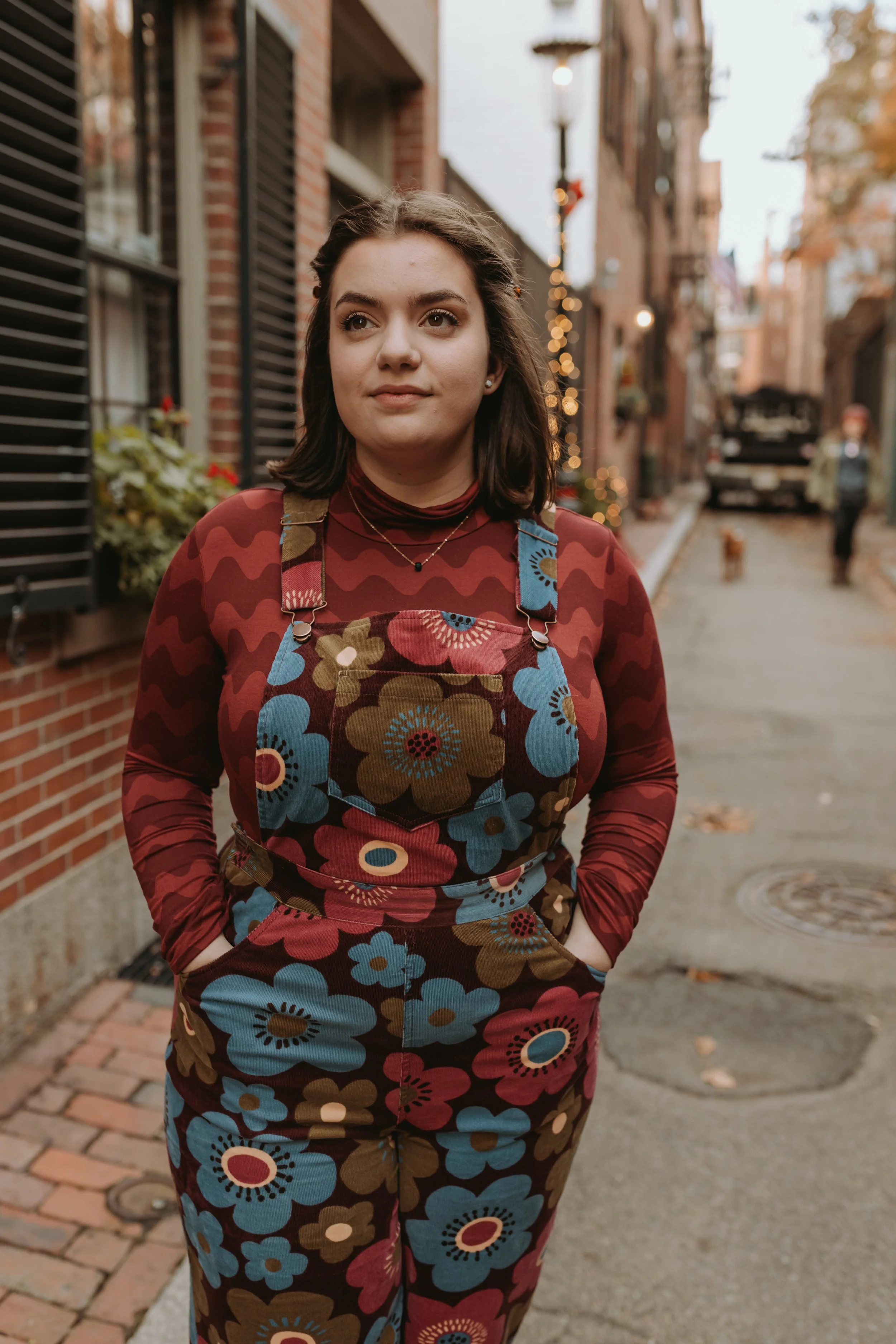 A young woman with dark brown hair wearing a colorful floral apron and a red patterned top, with hands in pockets, standing on a brick sidewalk in an outdoor urban setting decorated with Christmas lights.