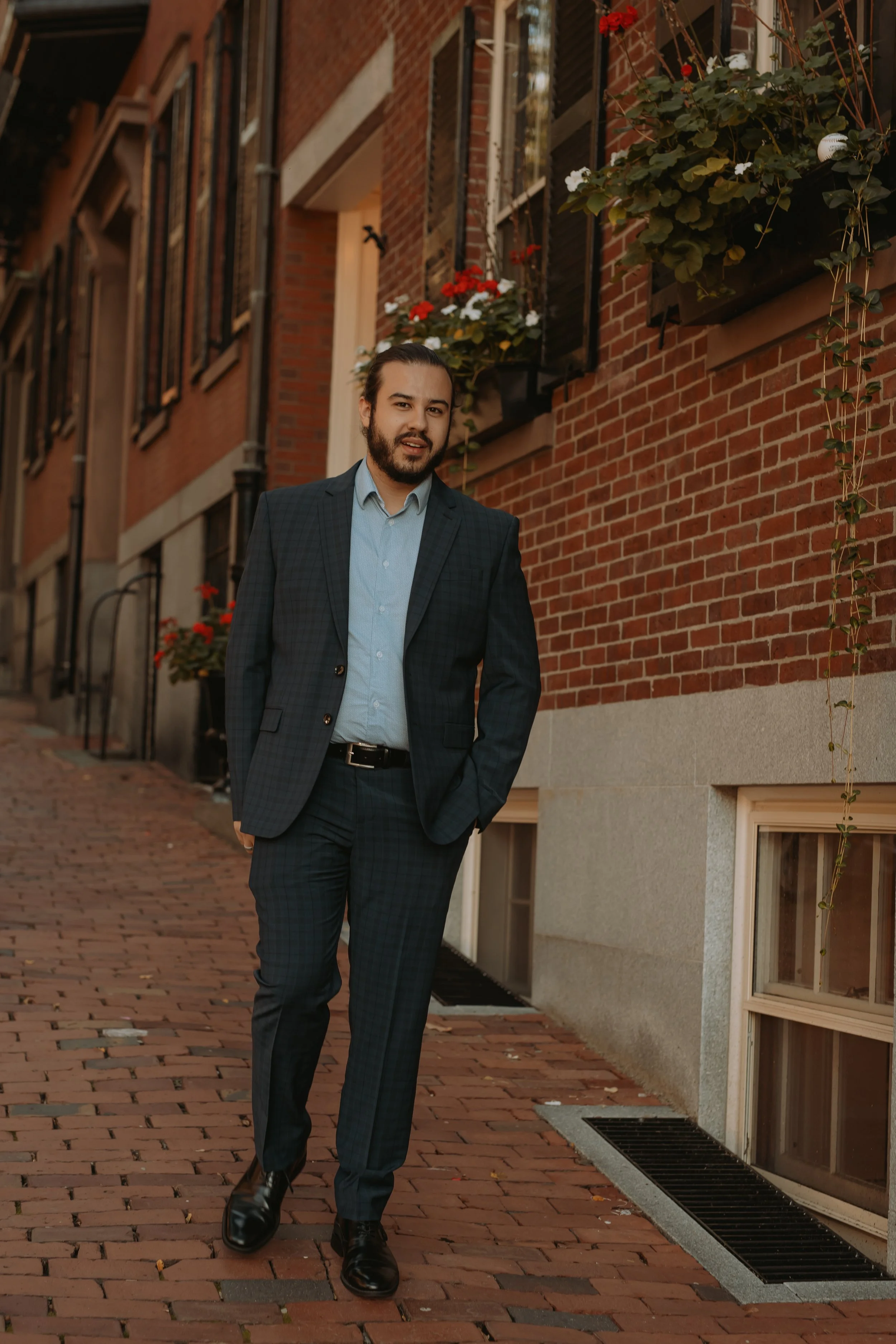 A well-dressed man walking on a brick sidewalk beside a brick building with flower planters and windows.