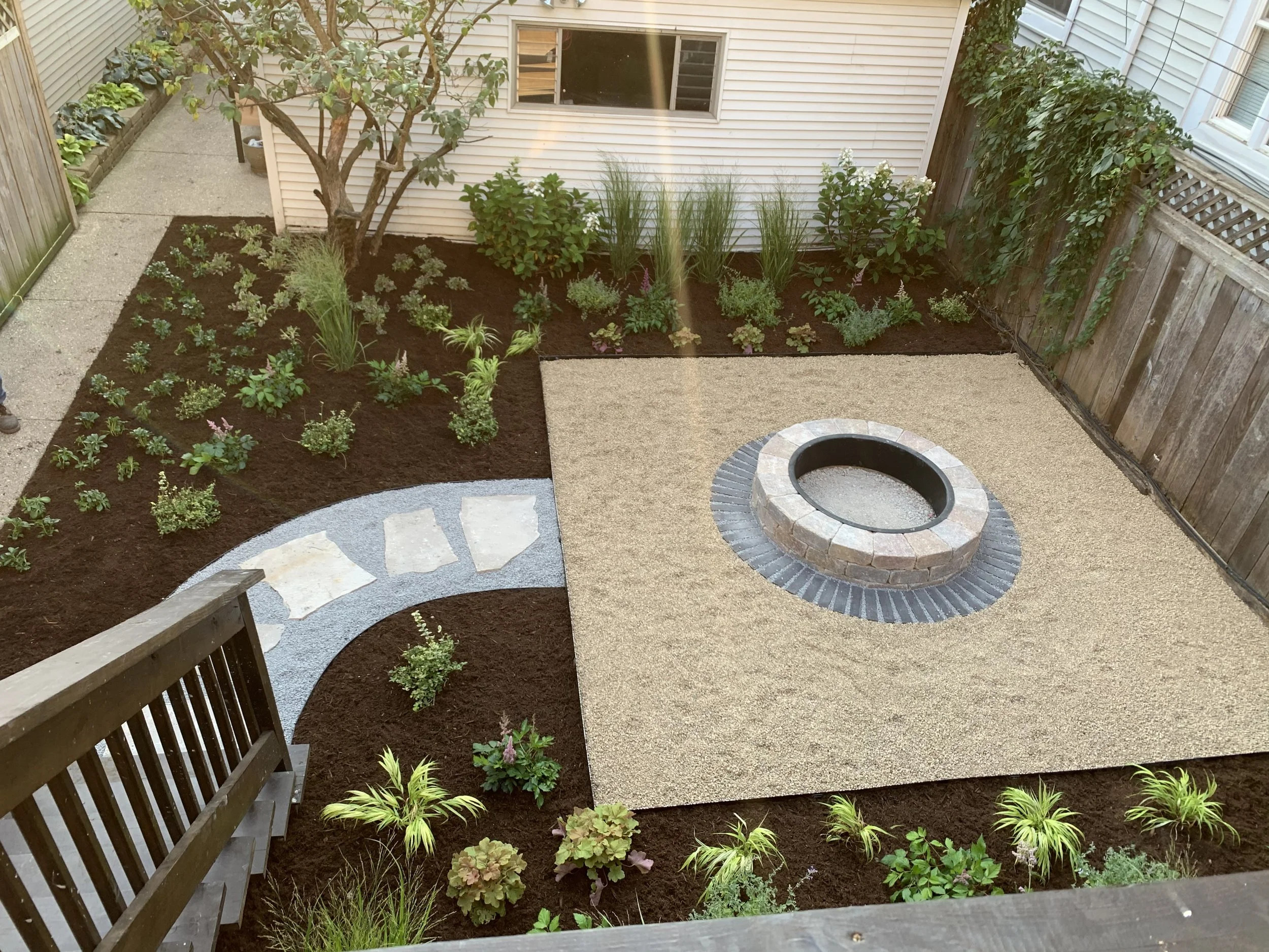 A backyard with a circular fire pit made of bricks in the center of a gravel area, surrounded by a patio area with sand-like material; a garden bed with diverse plants and mulch is along one side, with a small tree and a curved stone pathway