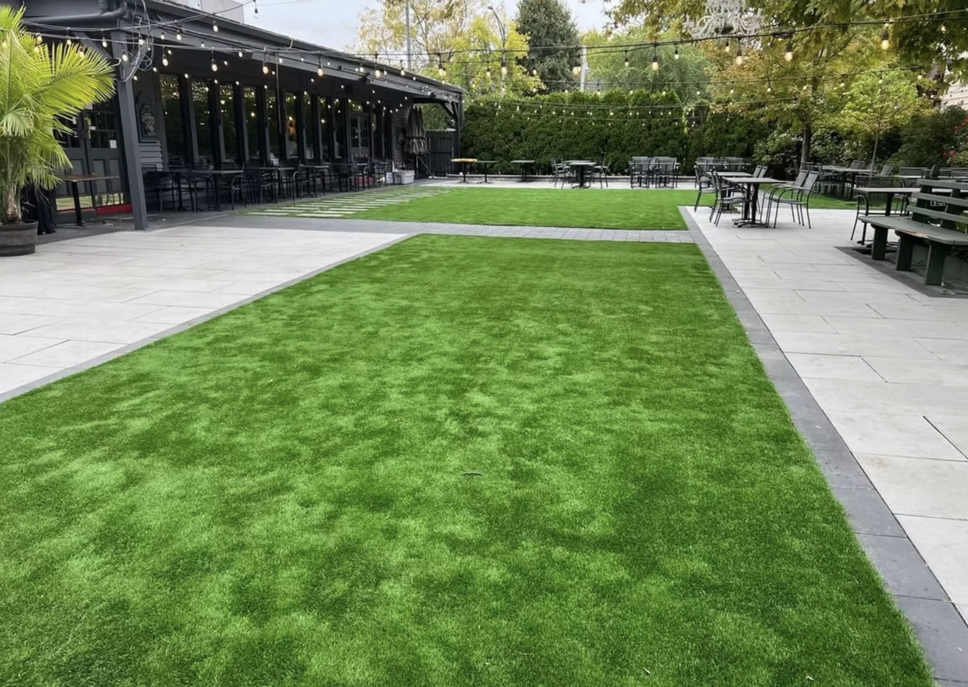 An outdoor patio with artificial grass, paved walkways, and string lights overhead, surrounding a black building with seating.