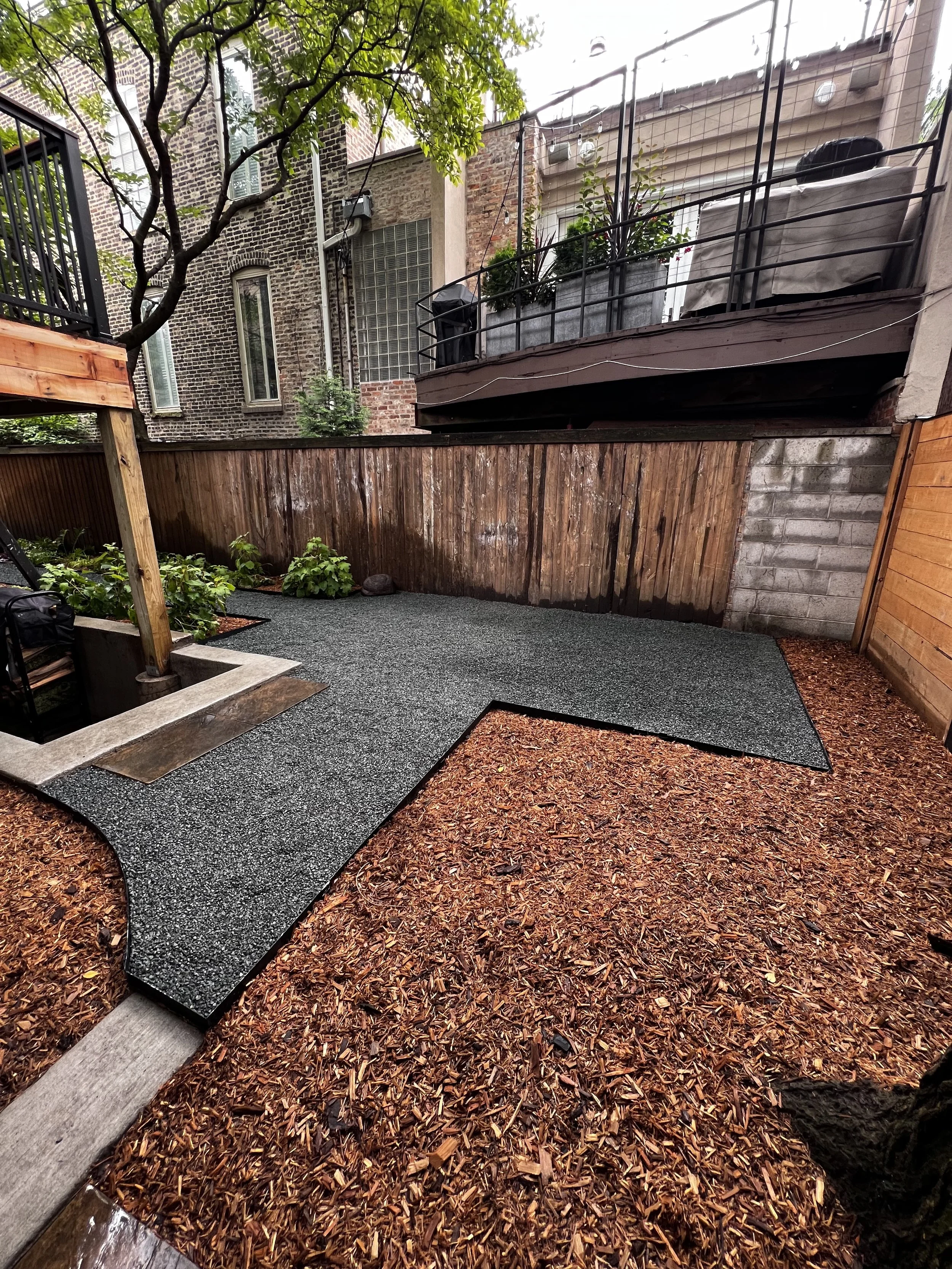 A small backyard with a gravel pathway, enclosed by wooden fences, with some potted plants and a tree arching over the space.