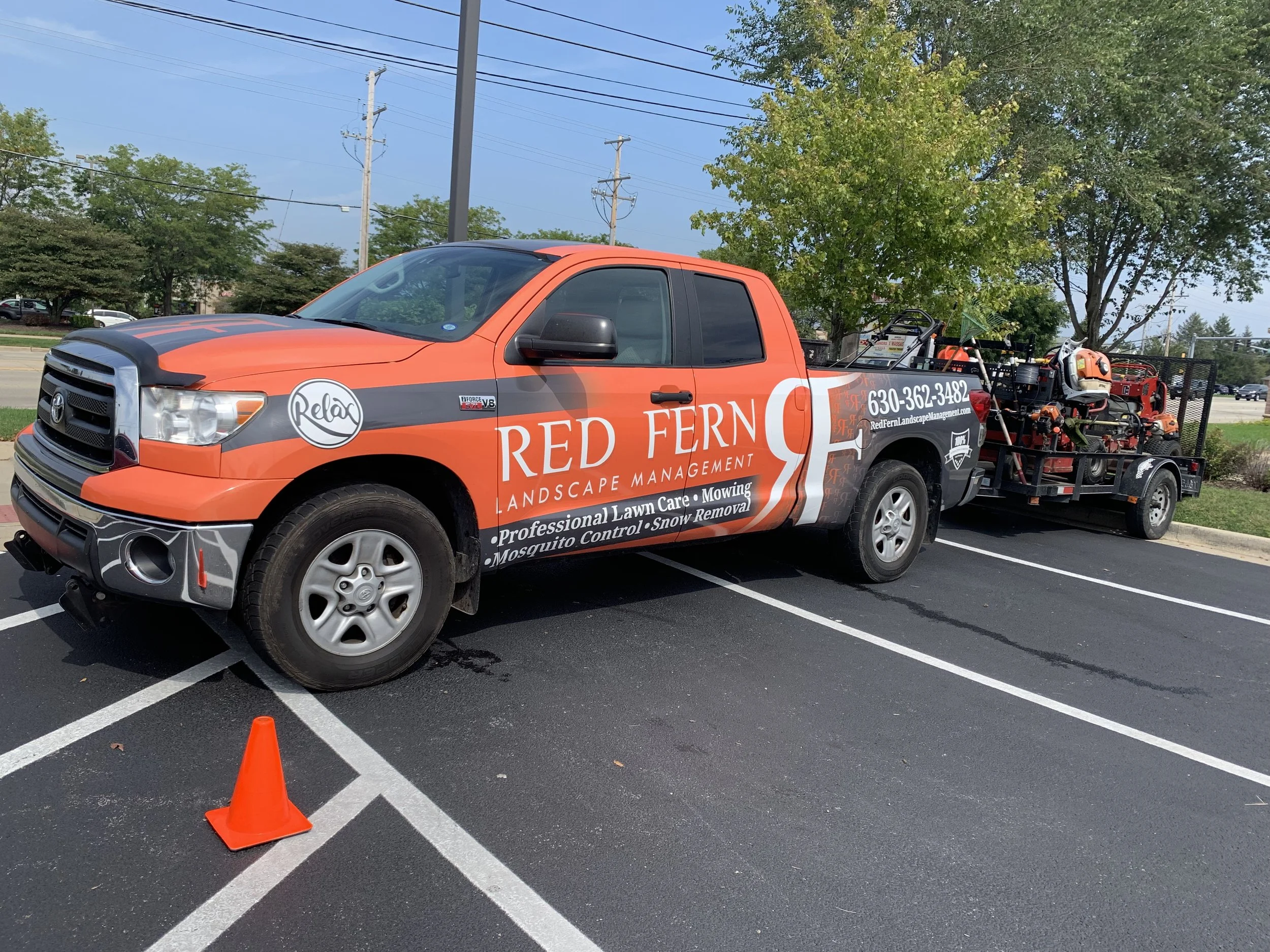 A red and black pickup truck with company branding parked in a parking lot, towing landscaping equipment on a trailer.