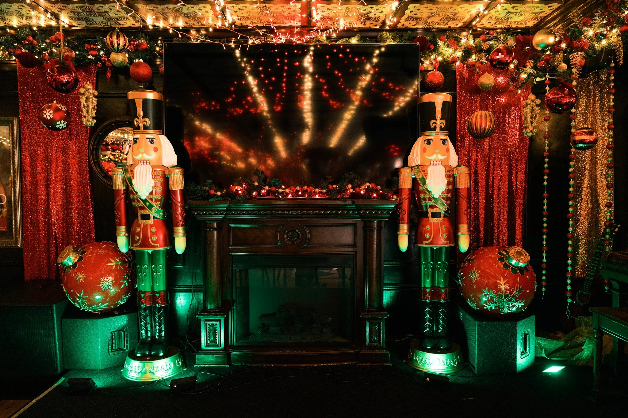 Christmas decorated interior with two nutcracker figurines, large red ornaments, red curtains, a fireplace, and string lights reflected in a large TV screen.