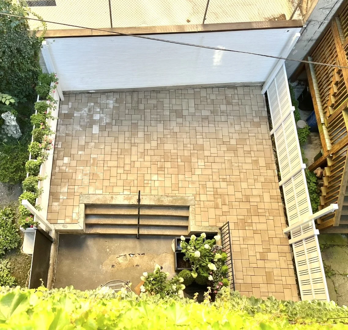 Top-down view of a backyard patio with tiled and concrete sections, stairs, plants, and a white fence.