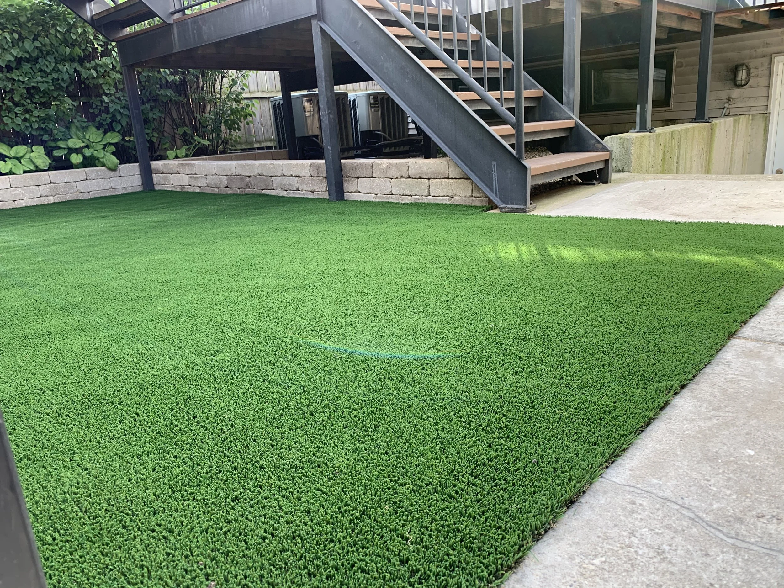 Artificial green turf lawn in a backyard under a staircase with a brick border and a concrete pathway.