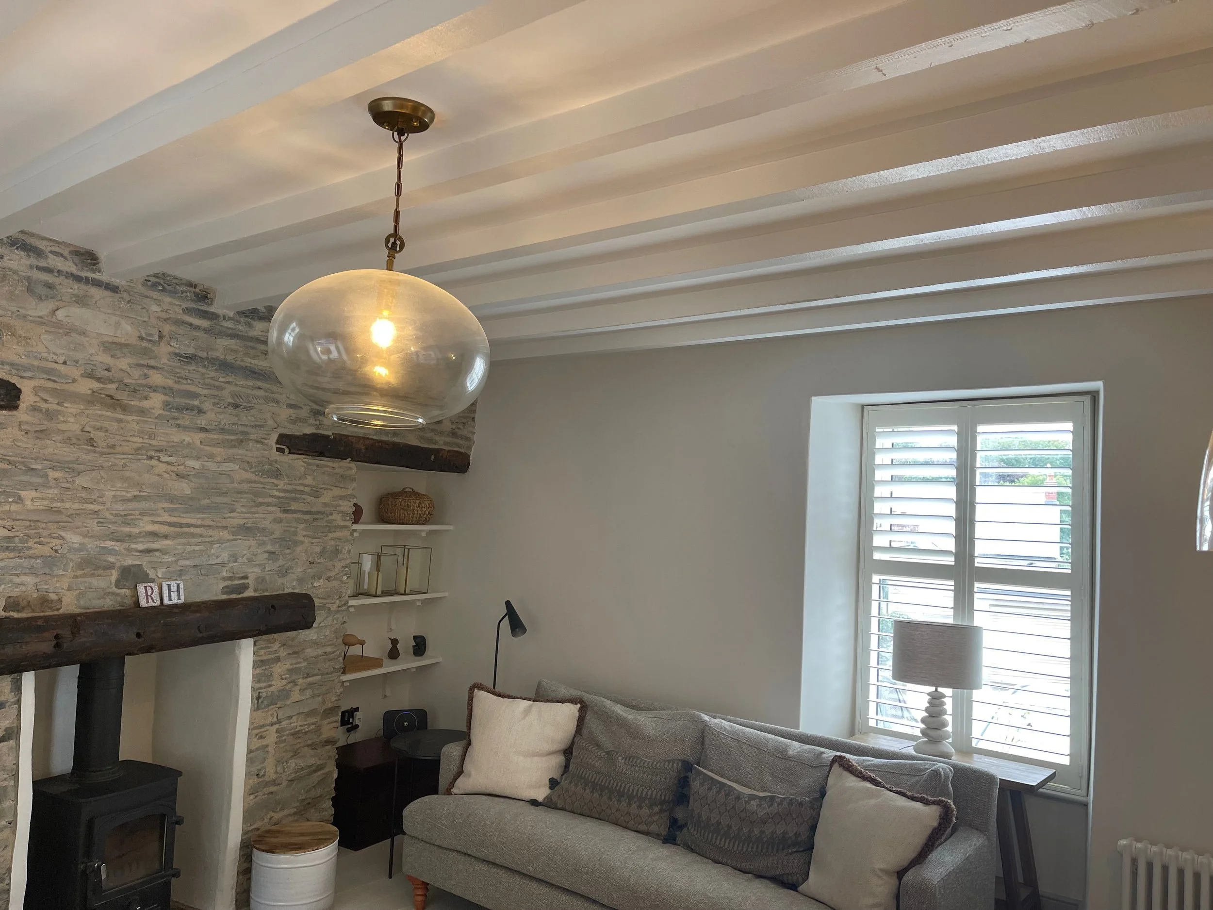 Living room with stone fireplace, wooden beam, hanging glass pendant light, beige sofa with pillows, window with white shutters, side table with lamp, and built-in shelves.