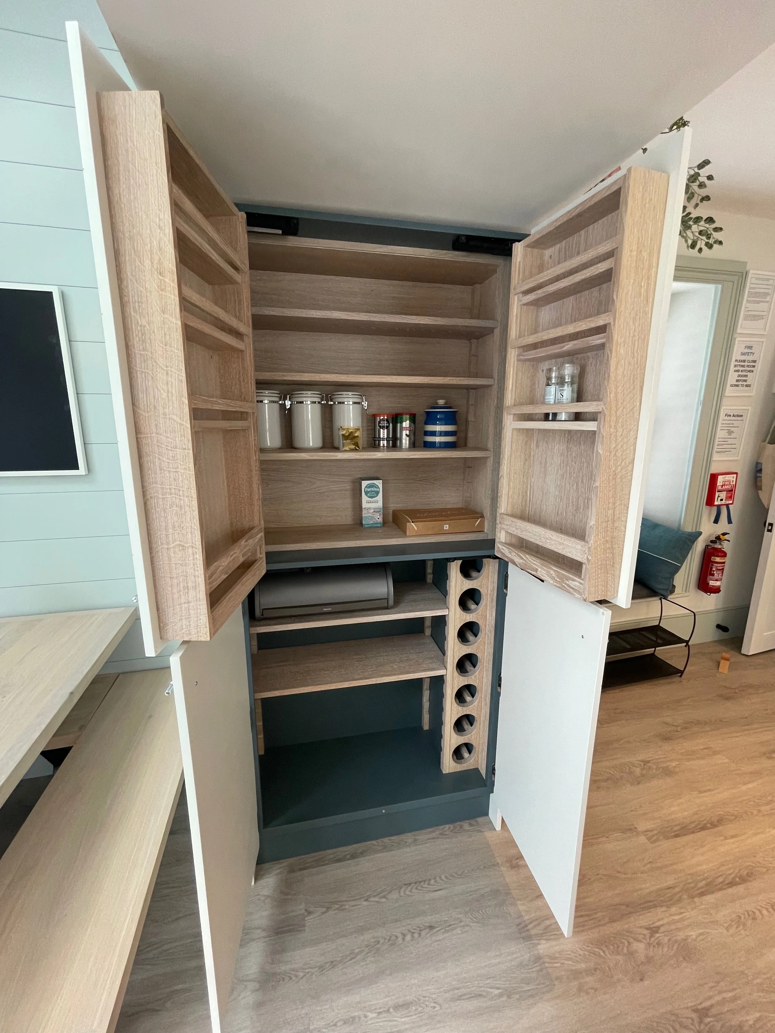 Open kitchen cabinet with wood and white doors, interior shelves, some jars, and a wine rack with circular holes.